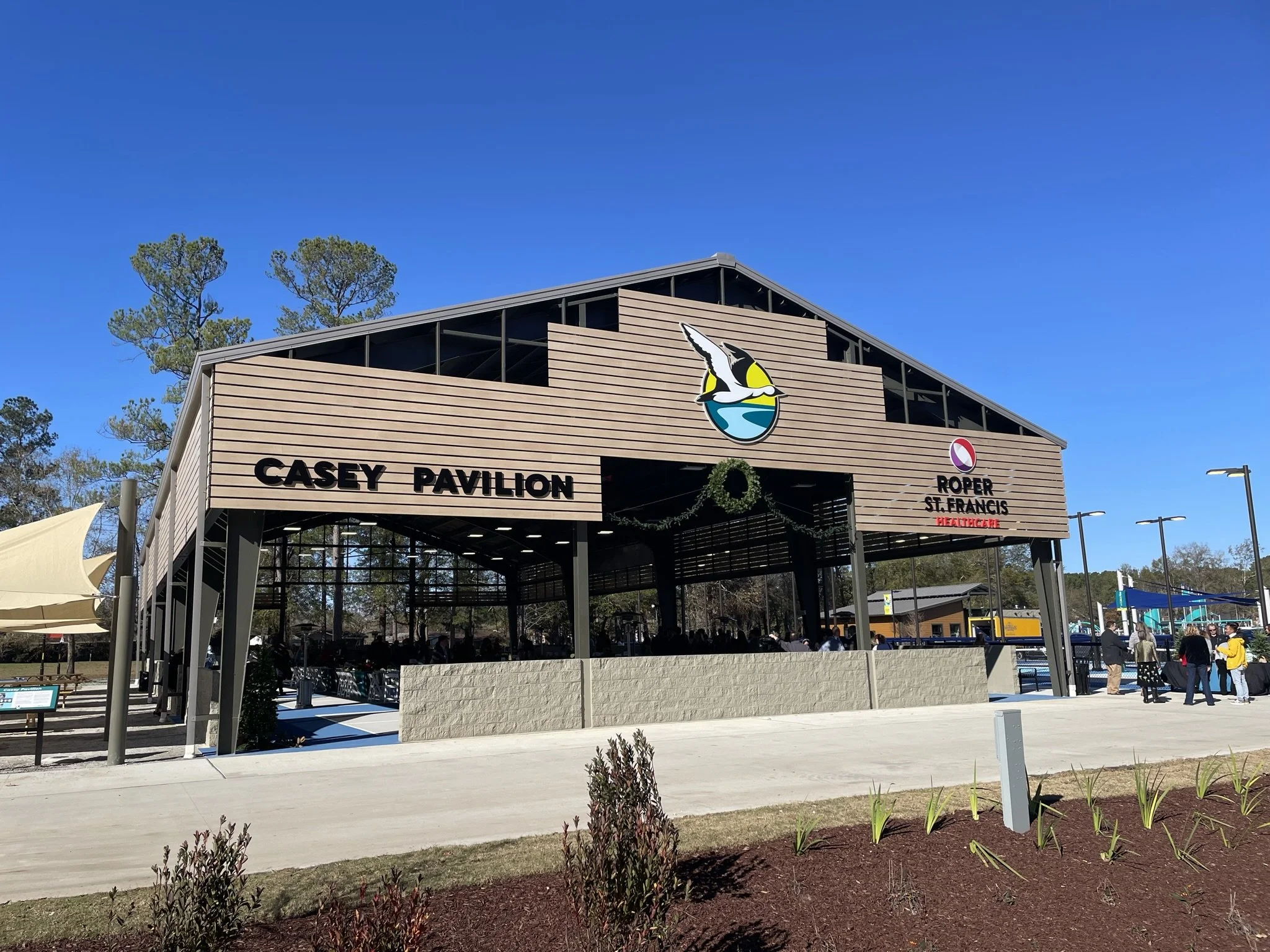 The Casey Pavilion at Roper St. Francis Healthcare is an open-air structure with a sloped roof and wooden siding, featuring a logo of a flying bird over a stylized landscape. People are gathered underneath, and the area is decorated with wreaths, indicating a festive occasion. The sky above is clear and blue, with trees in the background.