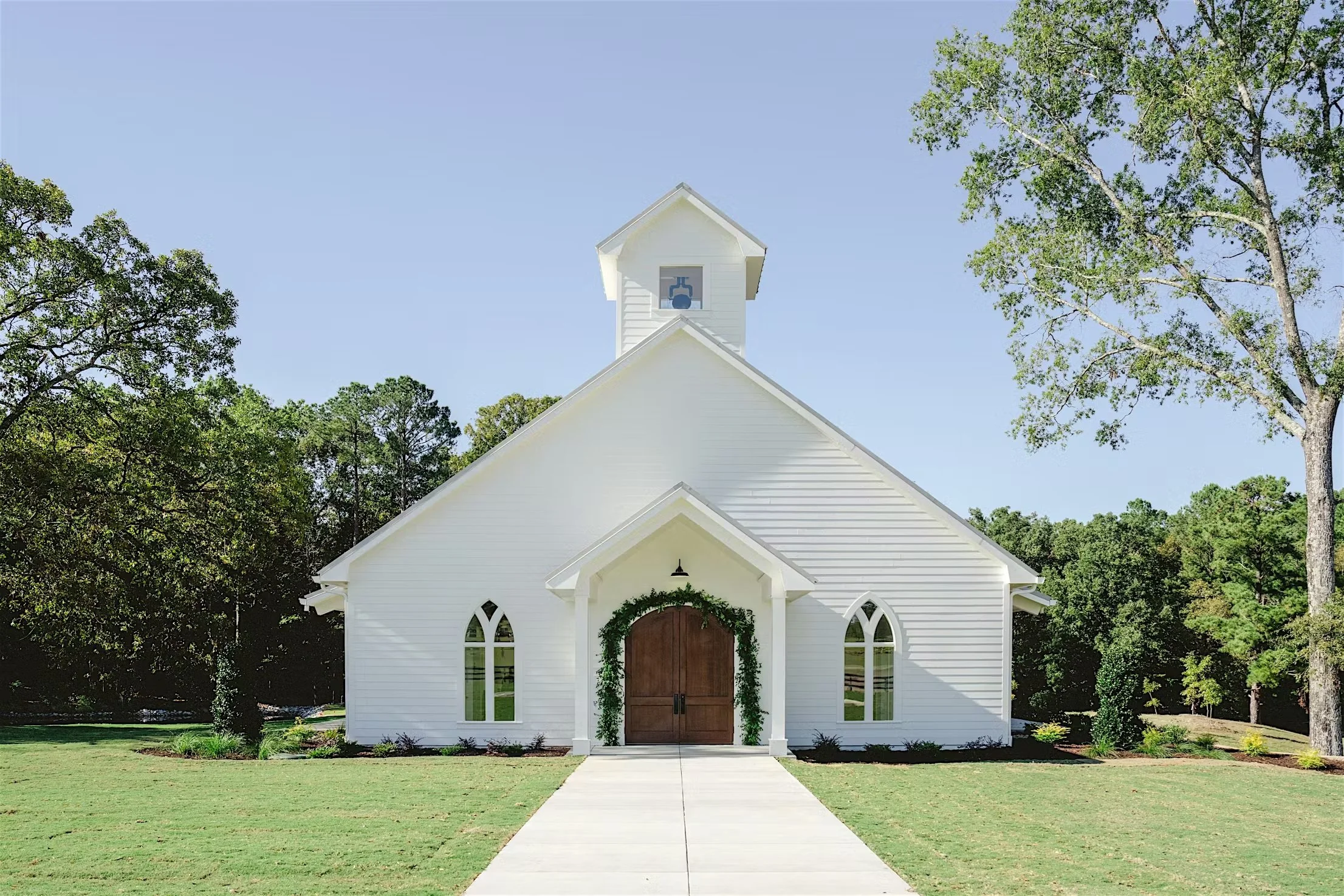 White church building with a peaked roof, arched windows, and a wooden door decorated with green foliage. Surrounded by green grass and tall trees, under a clear blue sky.