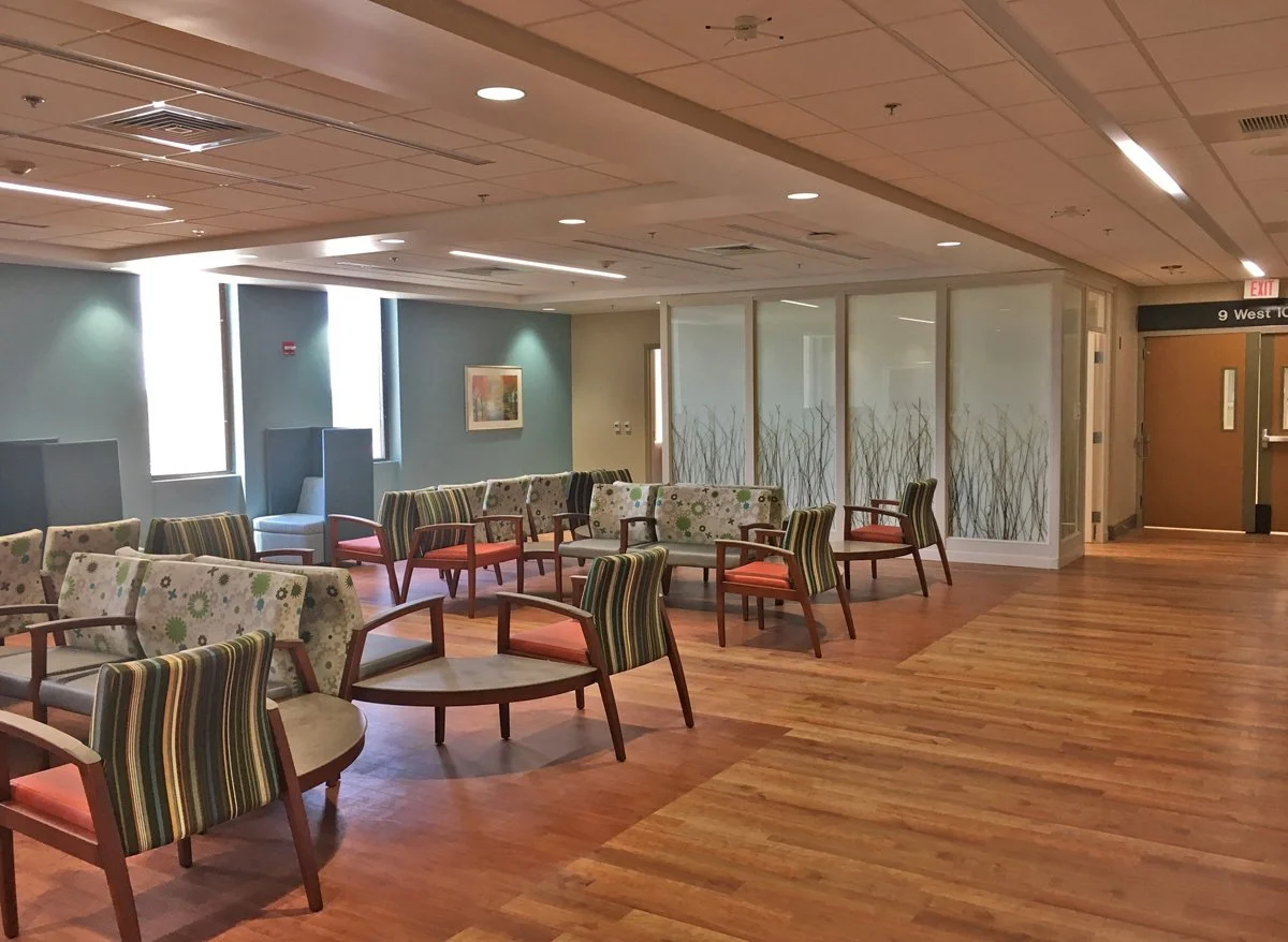 Empty waiting area with multiple chairs, some with striped fabric and some with floral patterns, wooden flooring, large windows, and a glass wall with decorative plant designs.