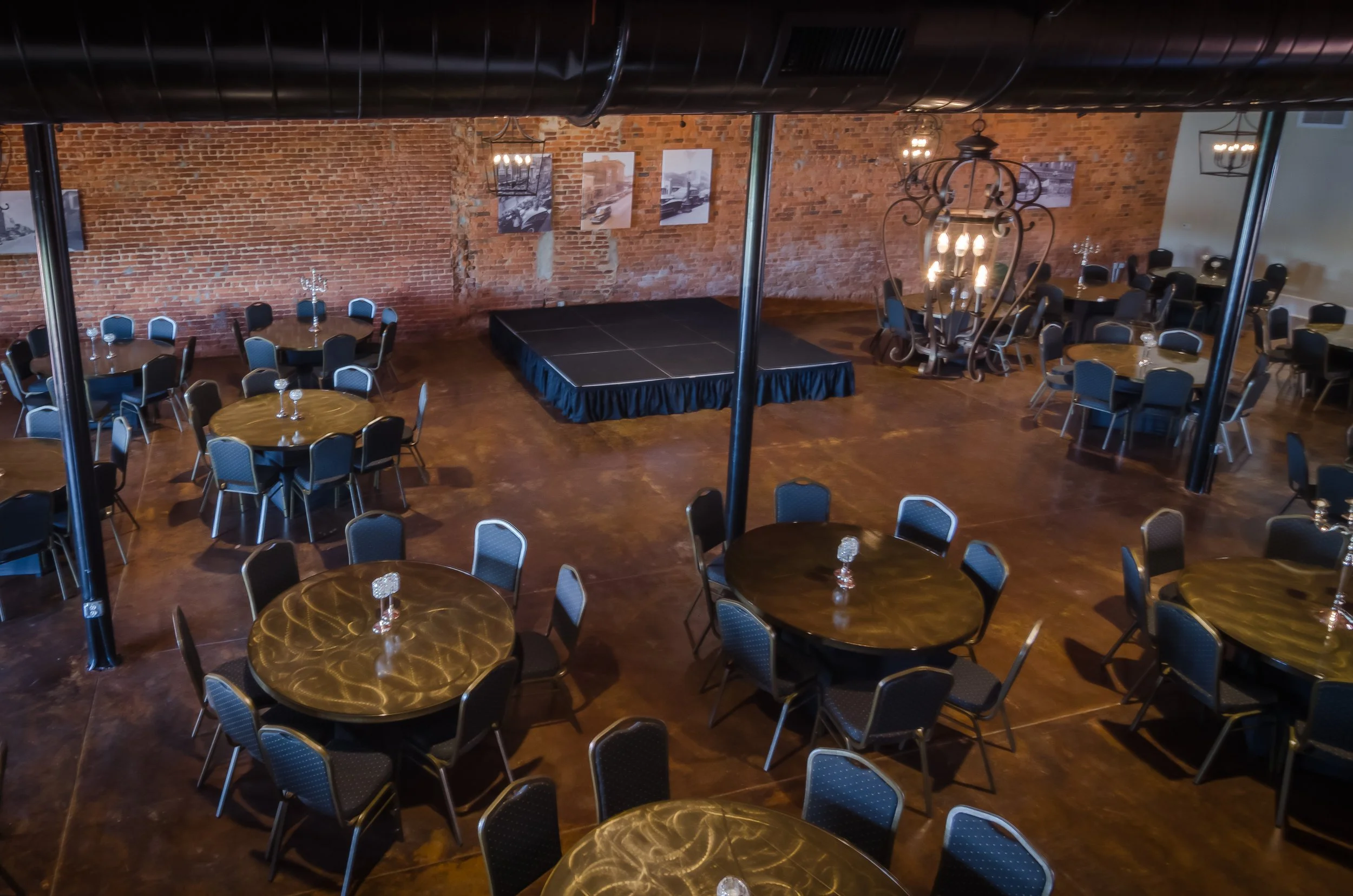 An event space with round tables covered with gold tablecloths and black chairs with blue cushions. There is a stage in the center with a black cover, and the walls are exposed red brick with some black and white pictures hanging. Chandeliers and wal
