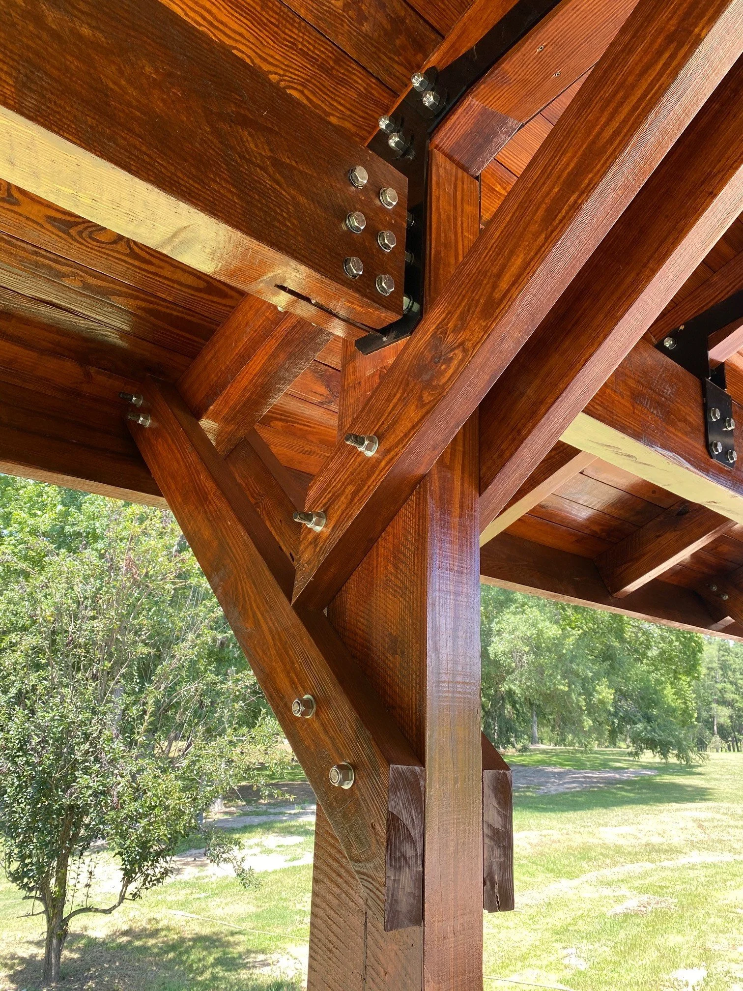 Close-up of a wooden structure, possibly a support beam or part of a pergola, with metal bolts and a black metal plate, outdoors with trees and grass in the background.