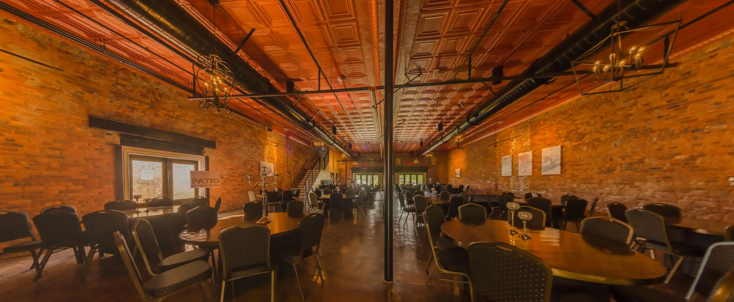 Interior of a rustic event space with exposed brick walls, wooden tables, and black chairs, illuminated by chandelier and wall-mounted lights.