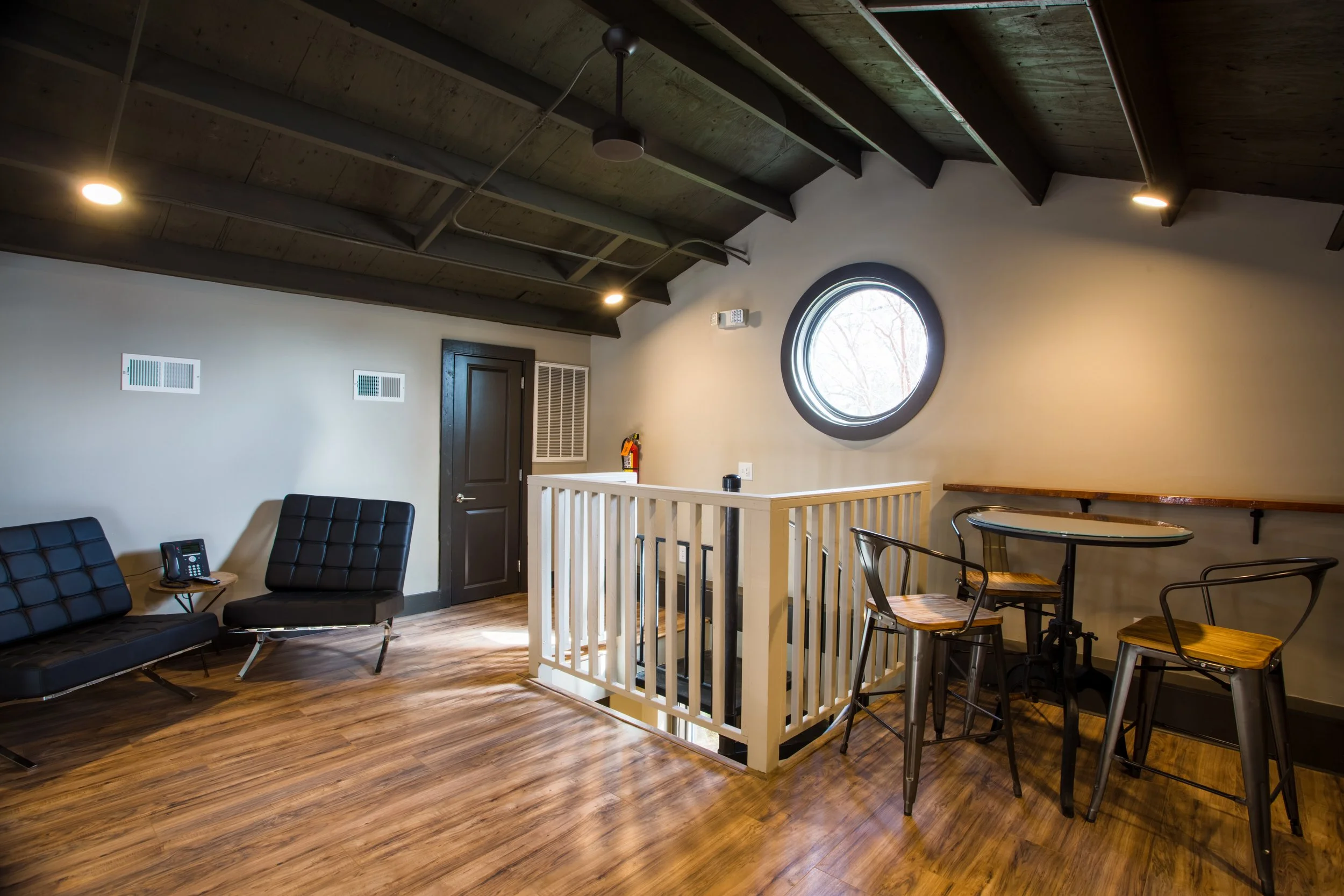 Interior of a modern room with wooden floors and a vaulted ceiling with exposed beams. There are black chairs, a small round table, and a white railing separating part of the space. A circular window shows trees outside, and wall-mounted heaters and 