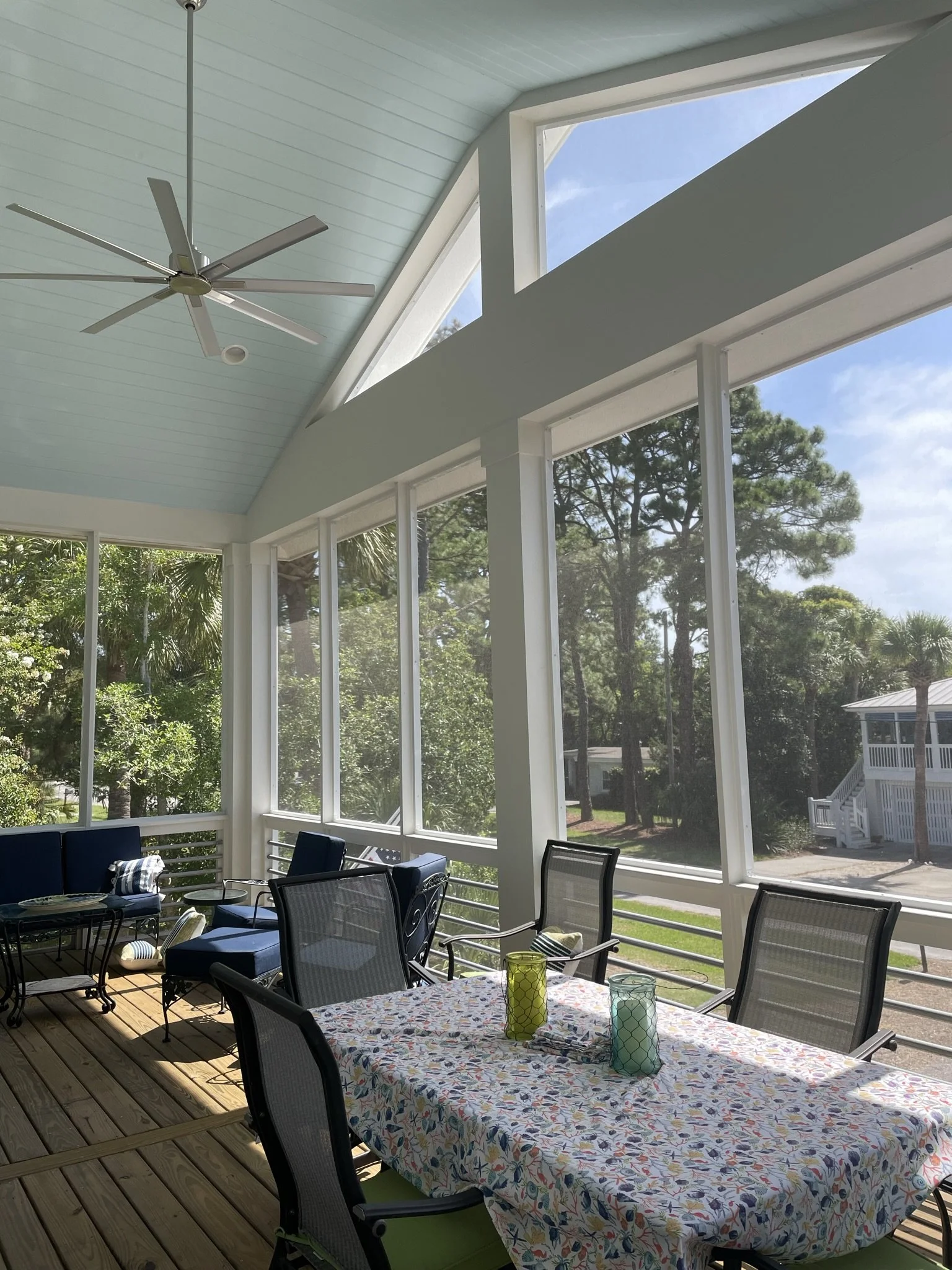 Sunroom with wooden floor, large windows, ceiling fan, outdoor scenery with trees, and a table covered with a colorful tablecloth and decorated with three blue, yellow, and green candles.