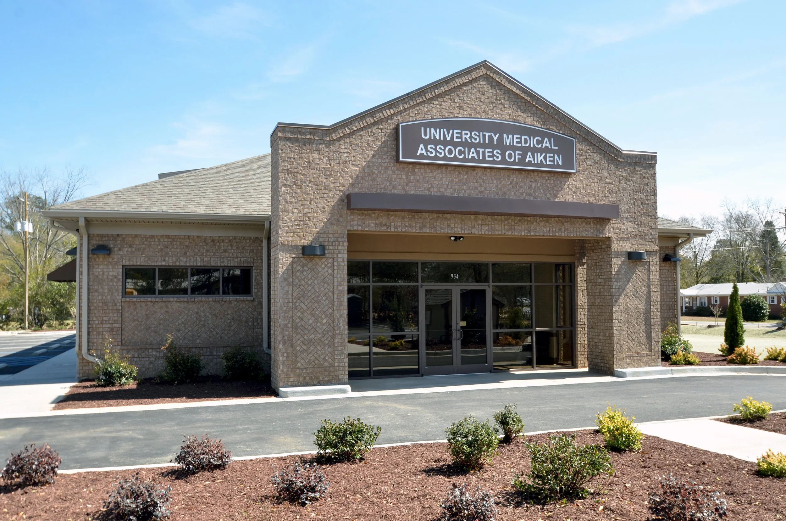 A brick building with a sign that reads 'University Medical Associates of Aiken' in an area with small bushes and plants in the foreground, and a bright blue sky in the background.