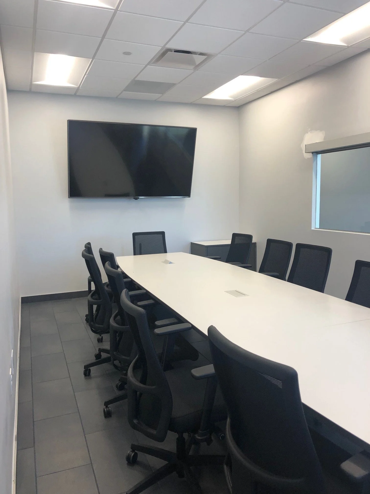 A conference room with a long white table and black office chairs. A large flat-screen TV is mounted on the wall, and there is a small window with a partially closed blind. The ceiling has fluorescent lights and an air vent.