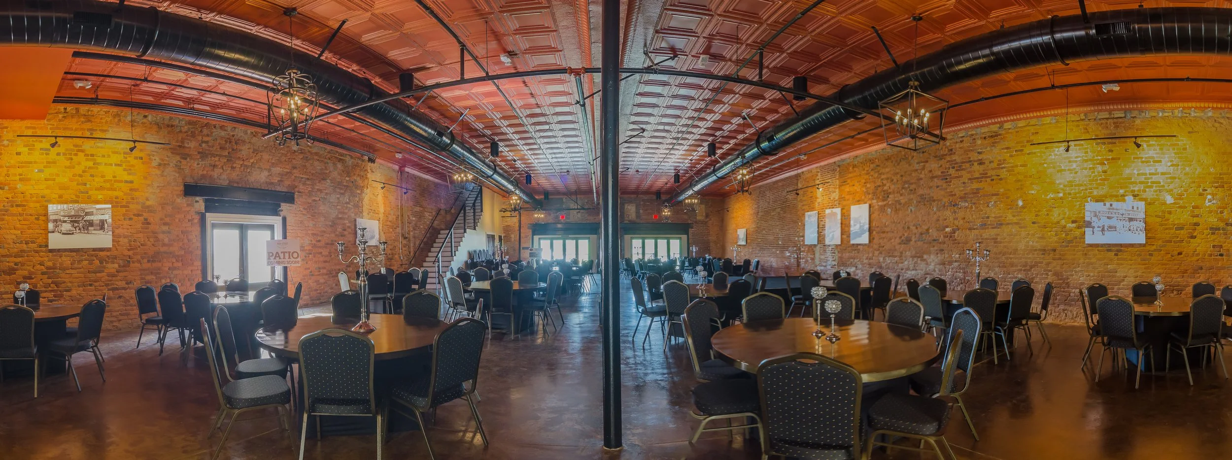 Interior of a banquet hall with round tables, chairs, candelabras, exposed brick walls, chandelier lighting, and a staircase at the back.
