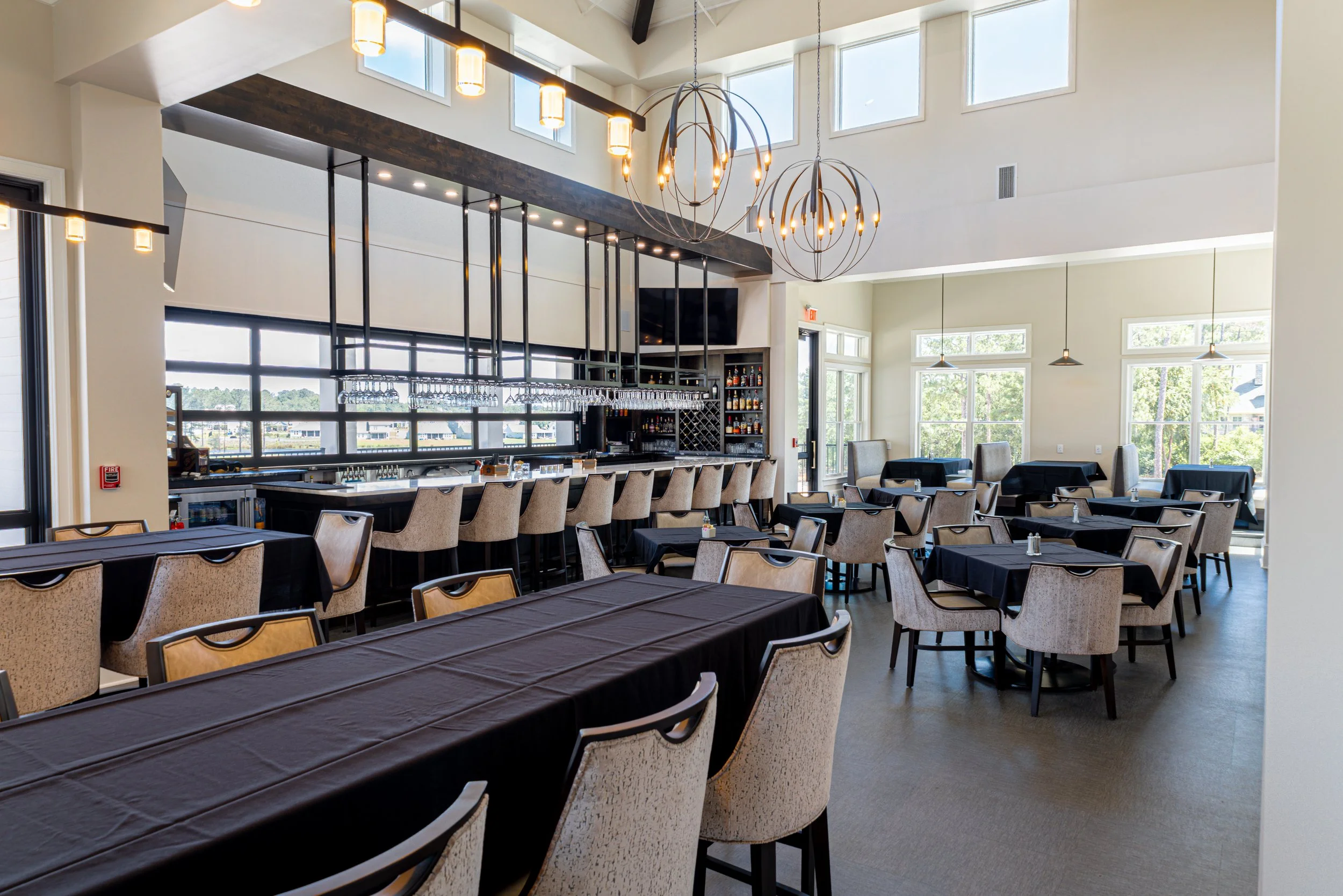 Interior of a modern restaurant or banquet hall with high ceilings, large windows, modern hanging light fixtures, a bar area with stools, tables with black tablecloths, and upholstered chairs.