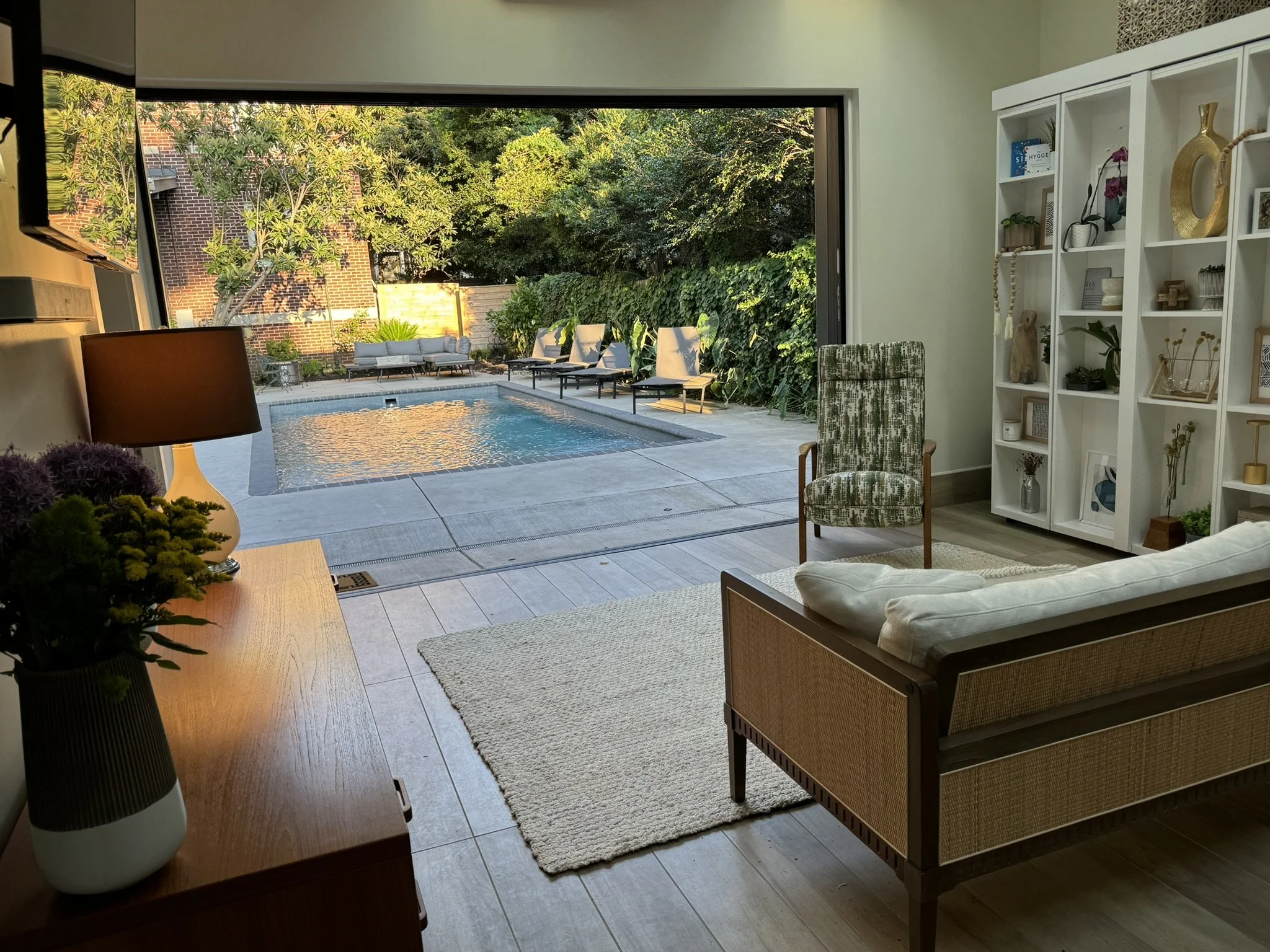 View from inside a house looking out through an open sliding door to a backyard with a swimming pool, outdoor chairs, and lush green trees and bushes.