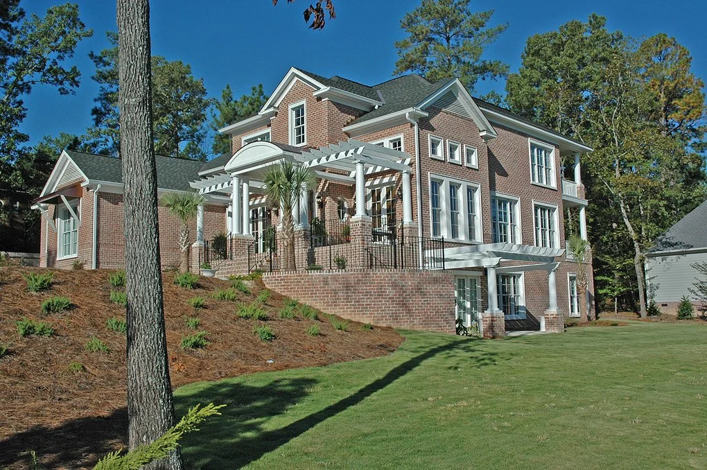 Large brick house with multiple balconies and a terraced backyard, surrounded by trees and grass, under a clear blue sky.
