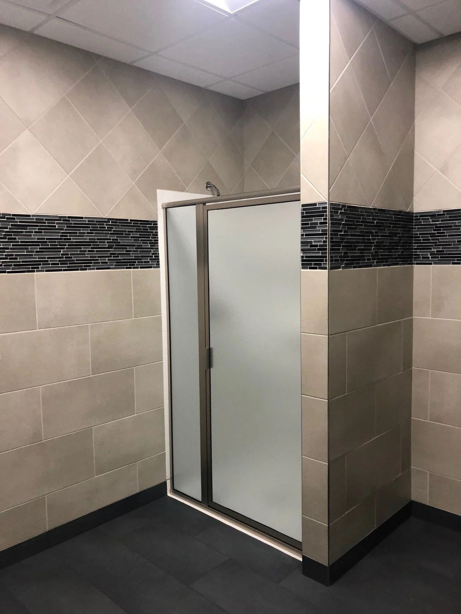 A public restroom with tiled walls and floor, featuring a locked paper towel dispenser, a black and gray mosaic tile accent band, and a partially open frosted glass door.