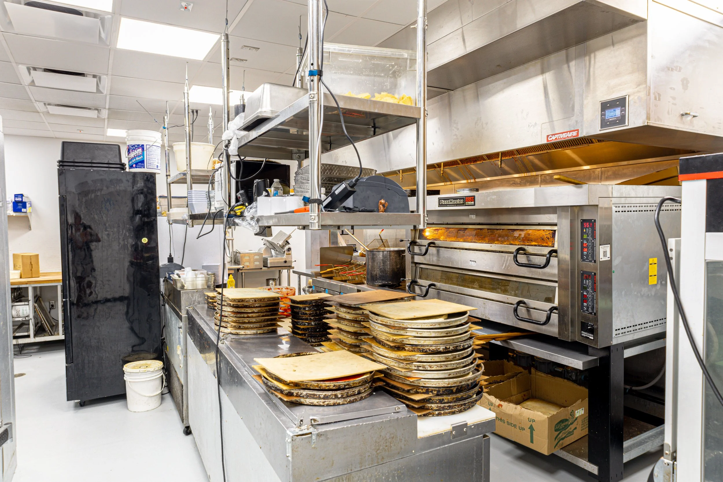 Commercial kitchen with stacked pizza pans and cooking equipment, including an oven, on stainless steel counters and shelves.