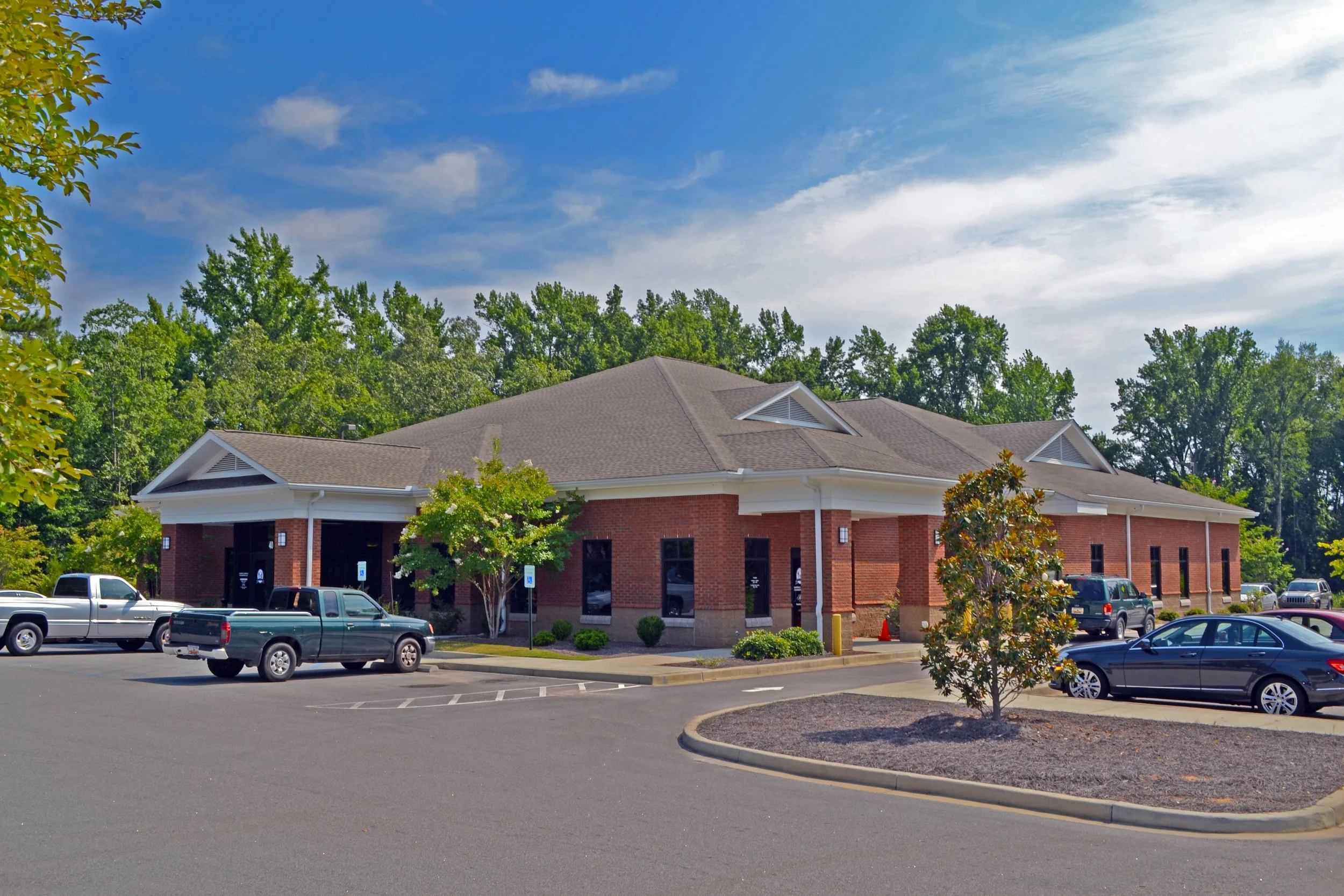 A brick building with a sloped roof and triangular vents on the sides, surrounded by trees and a parking lot filled with various cars, under a partly cloudy blue sky.