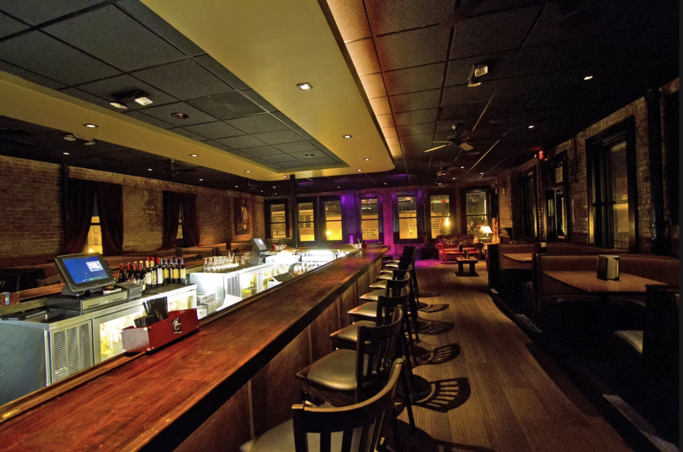 Interior of a dimly lit bar with a long wooden counter, some bar stools, and a brick wall. There are large windows with curtains, and ambient lighting with purple and warm tones.