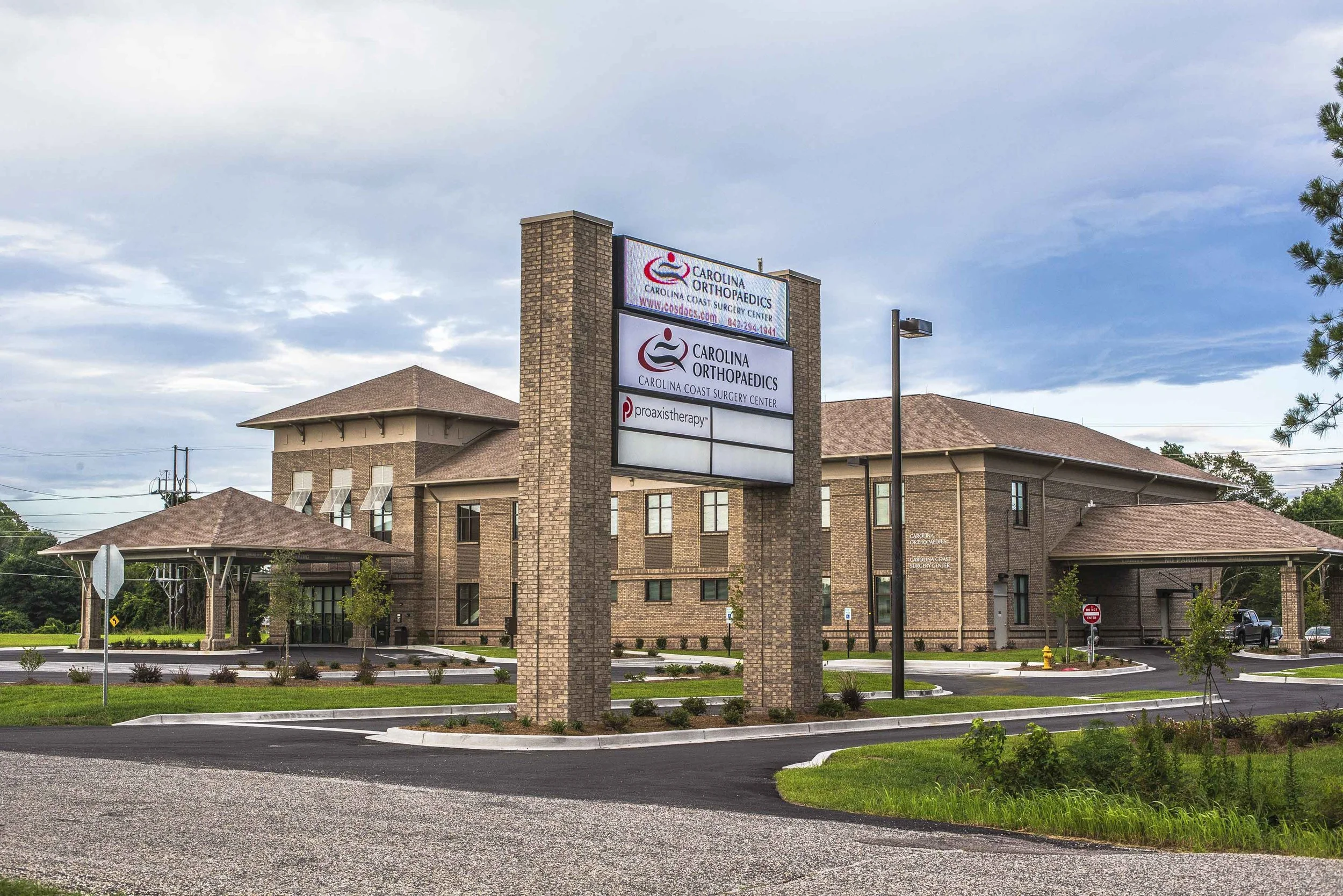 Exterior of a brick medical building with a tall sign displaying 'Carolina Orthopaedics' and 'Carolina Coast Surgery Center,' under a cloudy sky, with parking lot and landscaping.