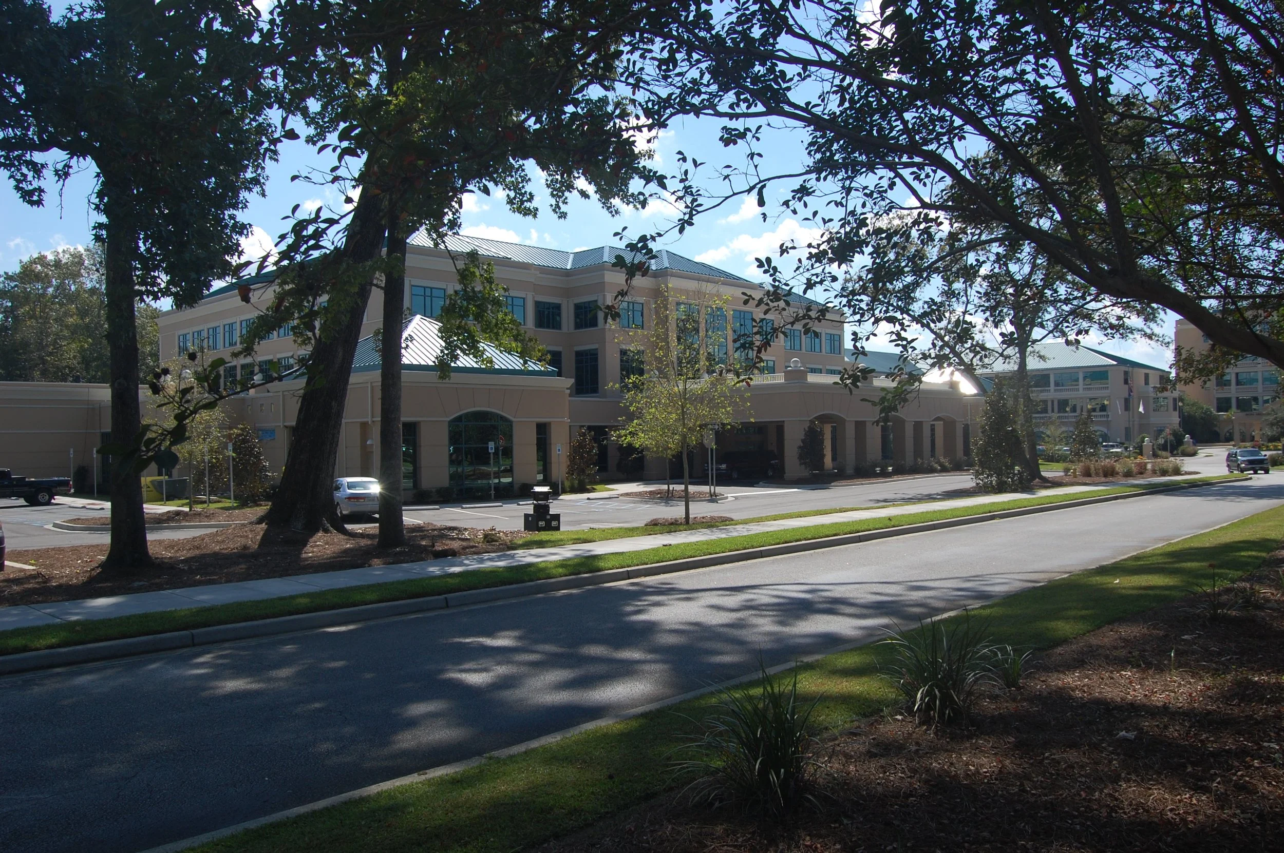 A large, multi-story building with a beige exterior and many windows, partially obscured by trees with green leaves, under a blue sky with some clouds. There are parked cars and a parking lot in front of the building.