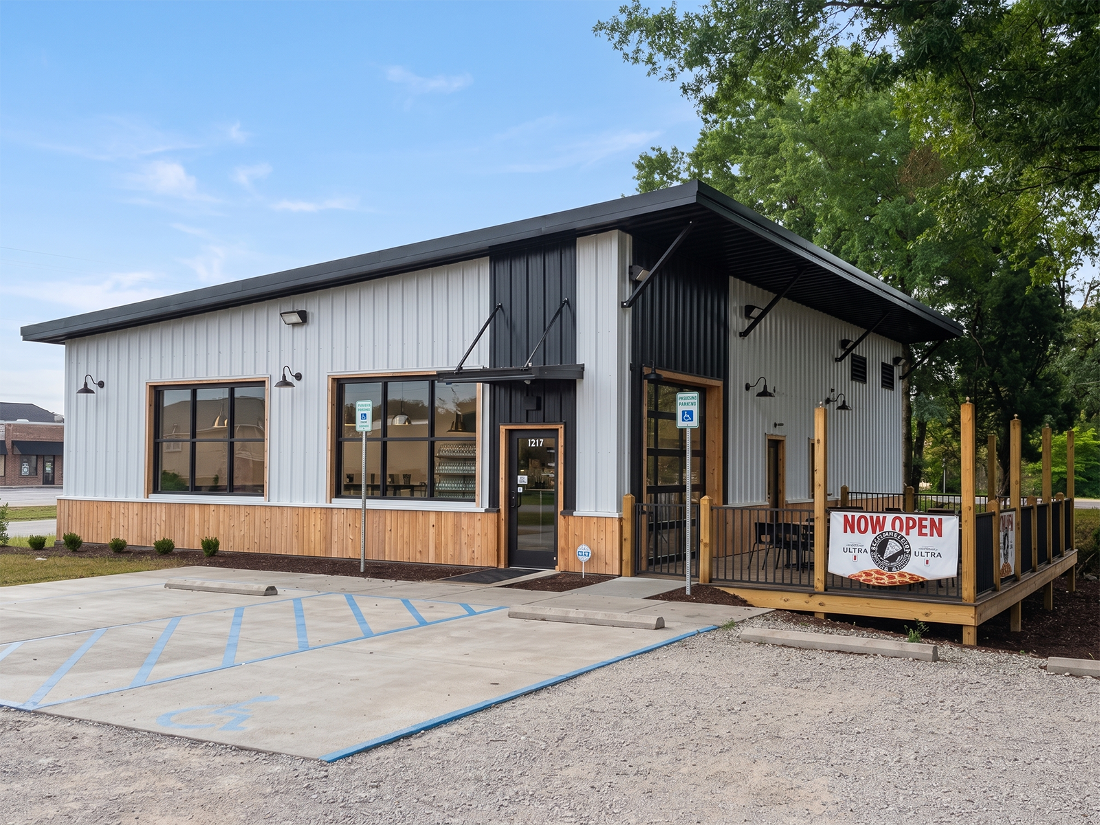 Modern commercial building with a metal exterior, large windows, and an outdoor patio area. A sign reads 'Now Open.' The parking lot shows designated spots for handicapped parking.