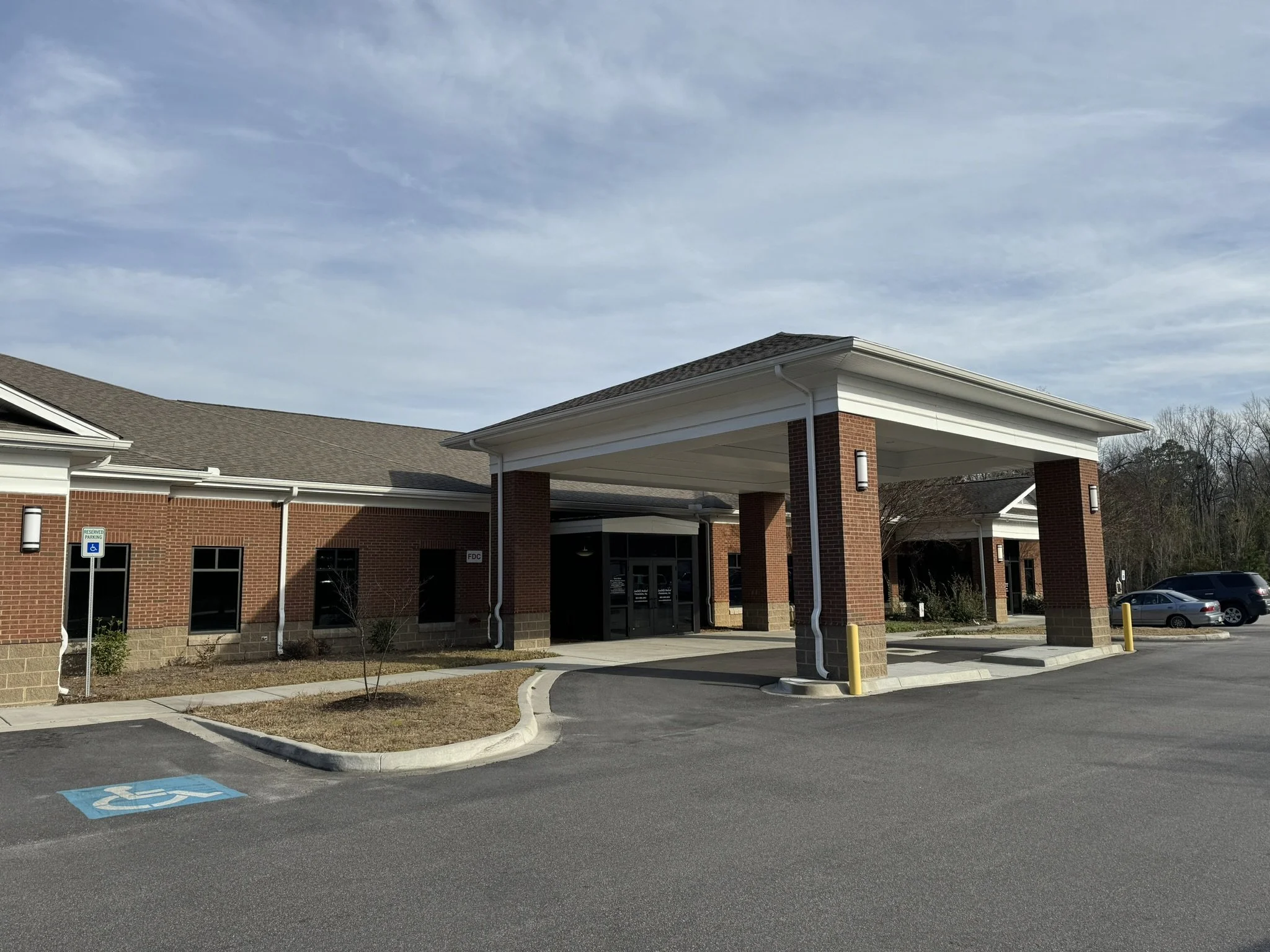 Exterior of a brick building with a covered entrance, parking lot with a handicap parking space, and a cloudy sky.