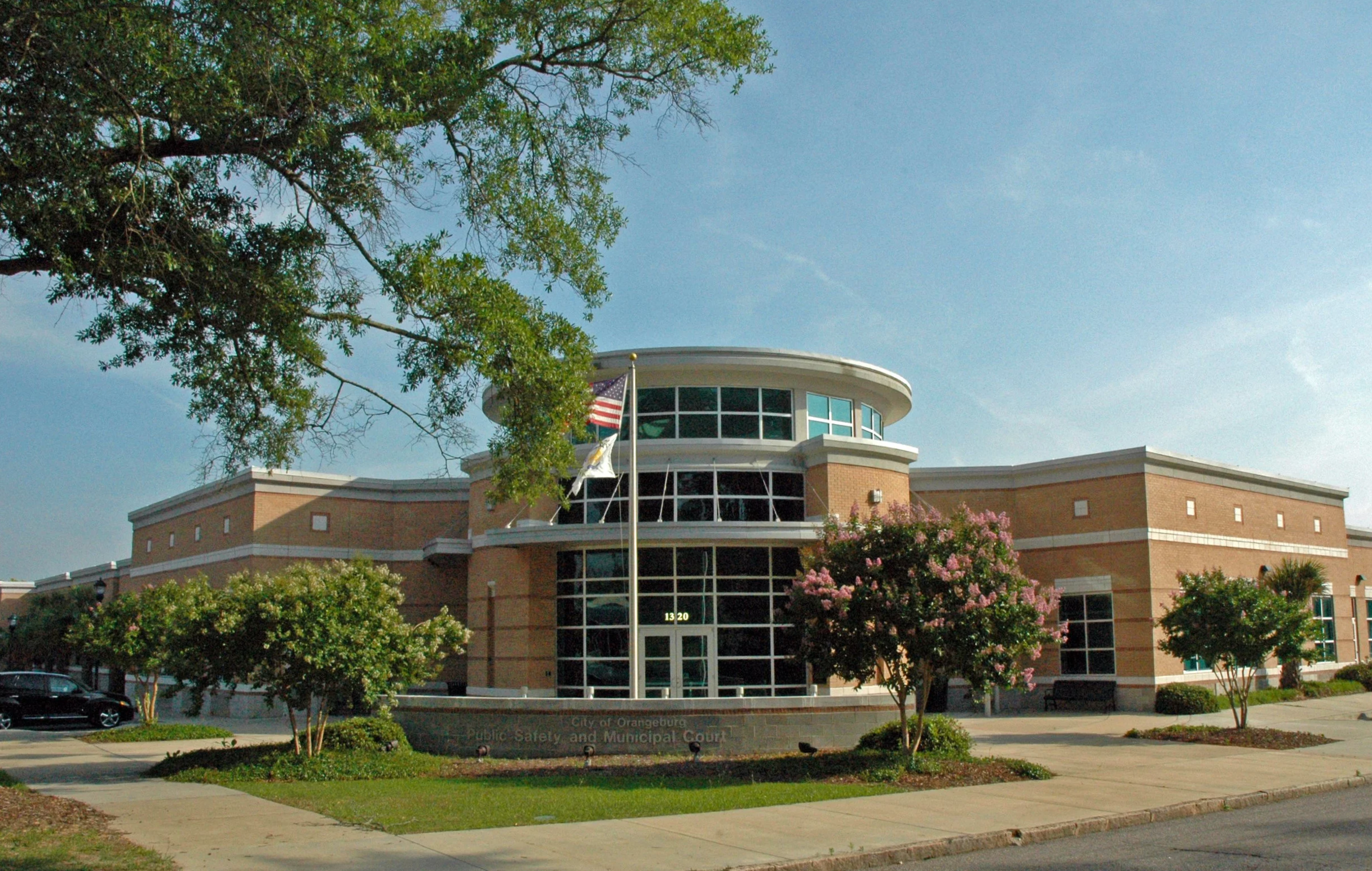 A brick municipal building with large glass windows, surrounded by trees and pink flowering bushes, with a flagpole and an American flag. Clear blue sky in the background.