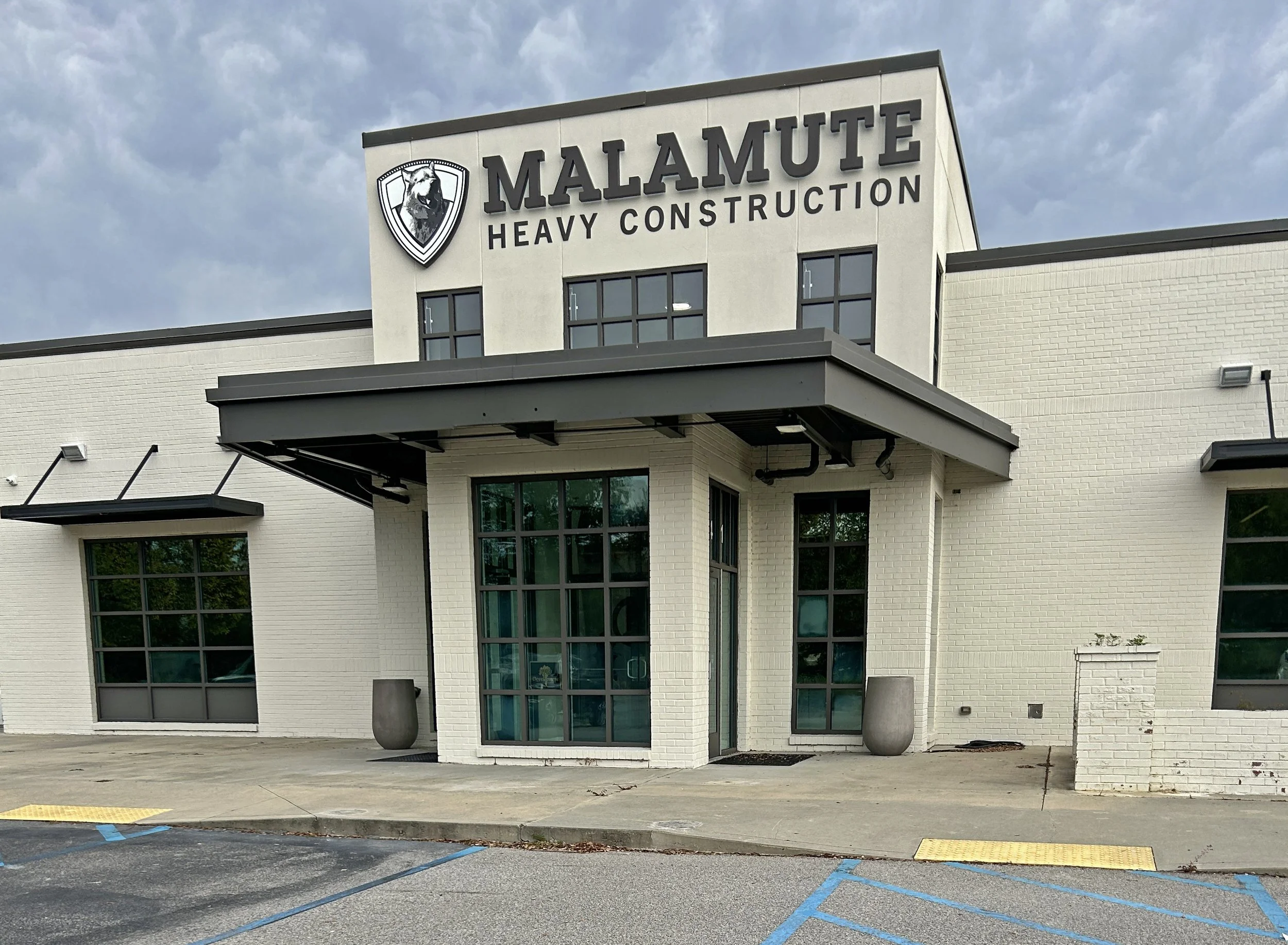 Exterior of a modern building with the sign 'Malamute Heavy Construction' and a wolf logo on the upper left corner. There are large windows and a small outdoor area with planters in front.
