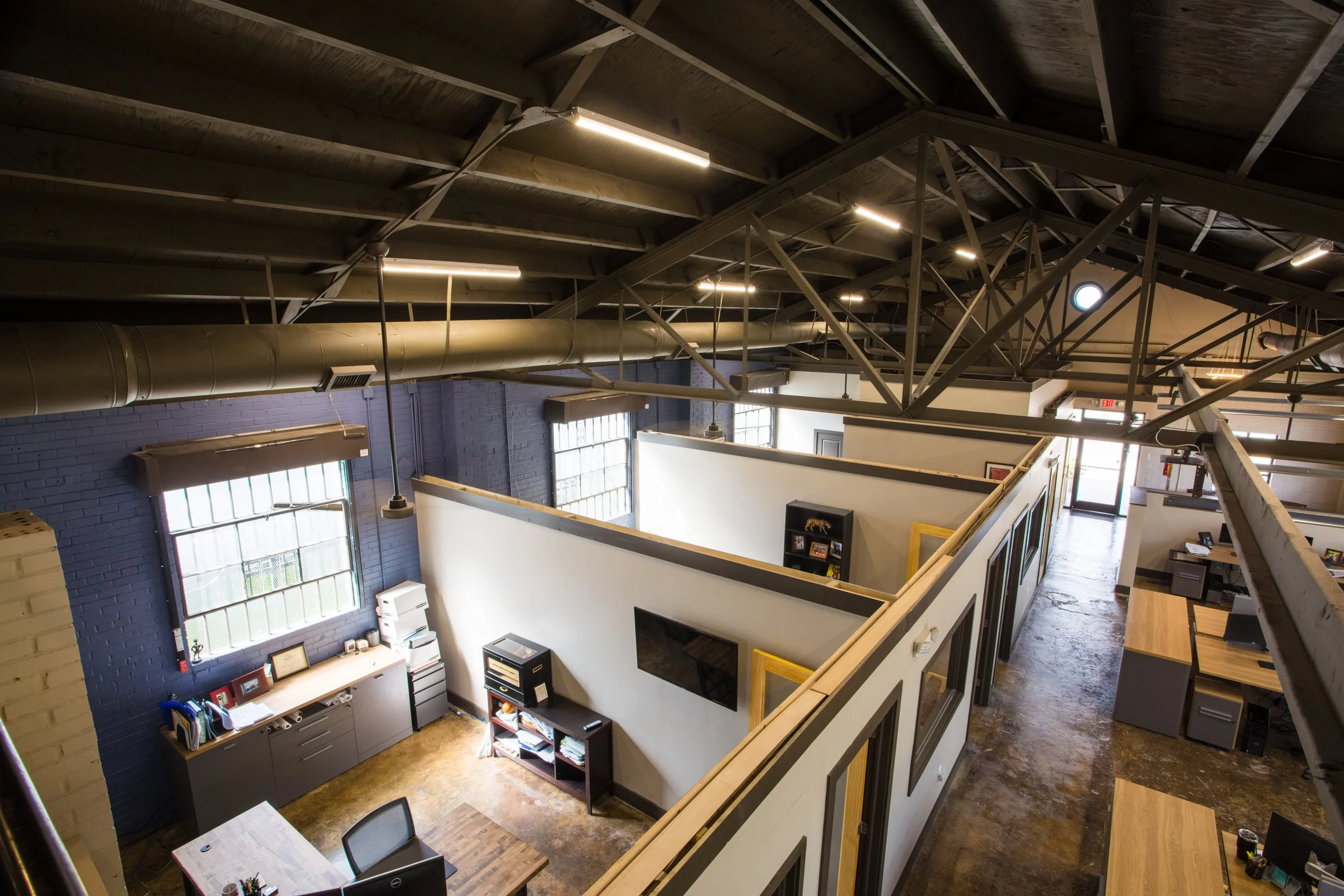 View of an industrial-style office space with exposed wooden ceiling beams, pipework, and modern lighting, featuring work stations, a blue brick wall, and large windows.