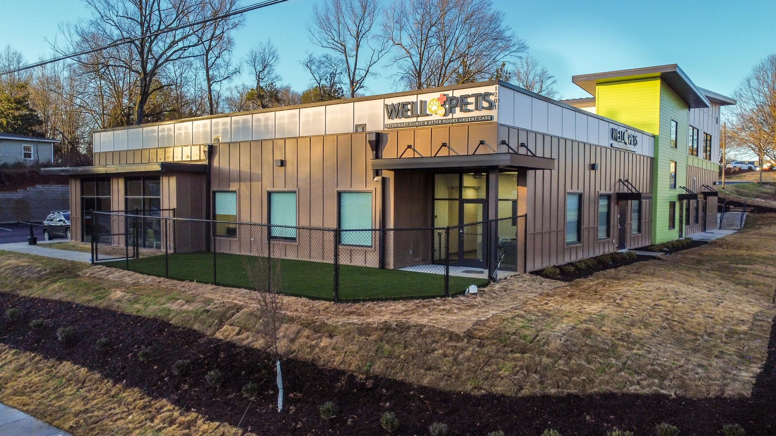 Modern veterinary clinic building with signage for Well & Pets, featuring a two-tone exterior with brown and bright green siding, large windows, and a fenced yard, set against a backdrop of leafless trees and a clear blue sky.