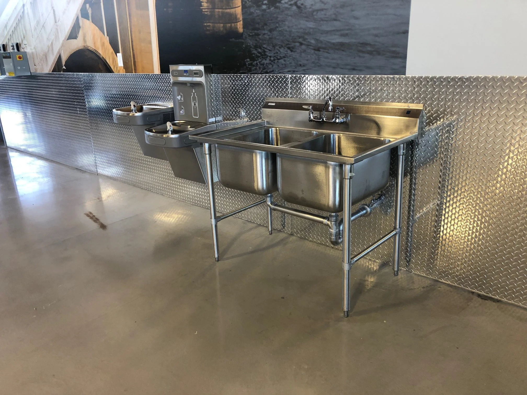 A stainless steel double sink with a tap is installed against a diamondplate metal wall, with a water fountain and some signs nearby, in what appears to be a public restroom or wash area.