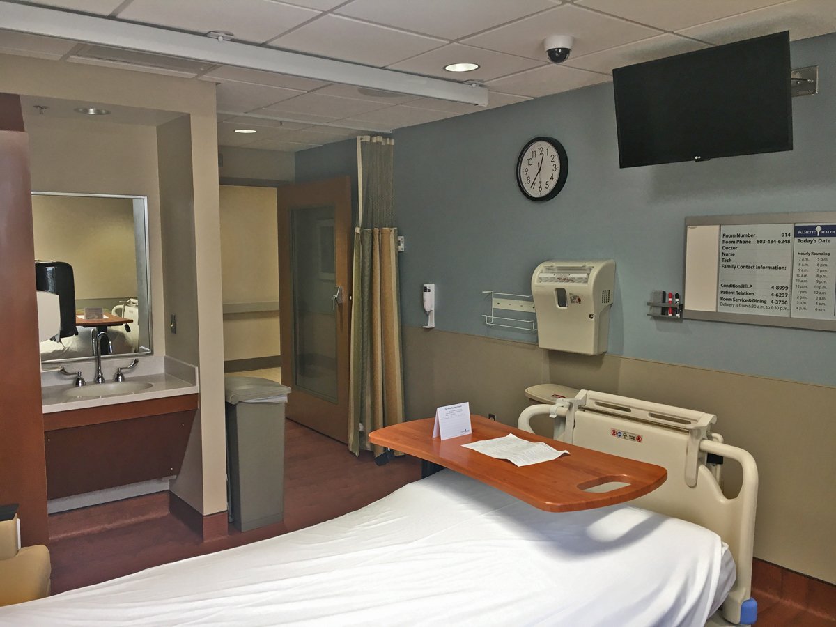 Hospital room with bed, bedside table, wall clock, television, whiteboard, and medical equipment.