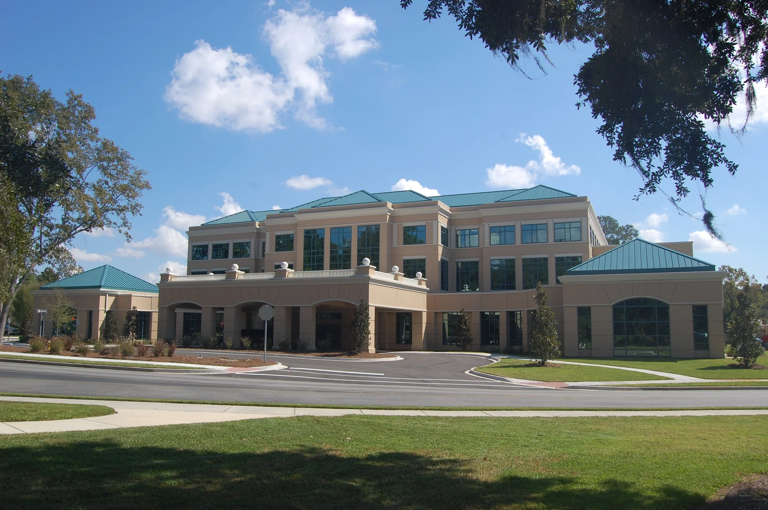 A large beige building with multiple glass windows and a teal metal roof, surrounded by trees with green leaves, under a partly cloudy blue sky.