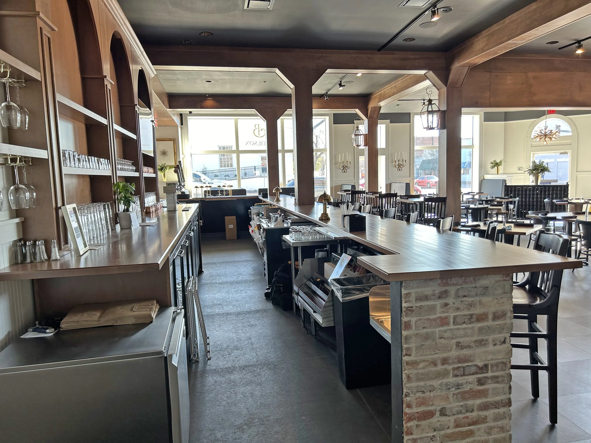 Interior of a restaurant with a long wooden bar counter, glassware on shelves, and tables and chairs near large windows allowing sunlight inside.
