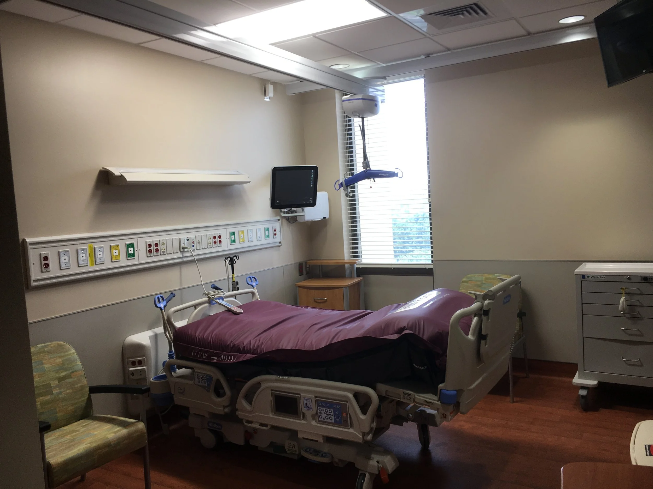 A hospital room with a bed and medical equipment, including a monitor, wall outlets, and medical devices, illuminated by natural light from a window.