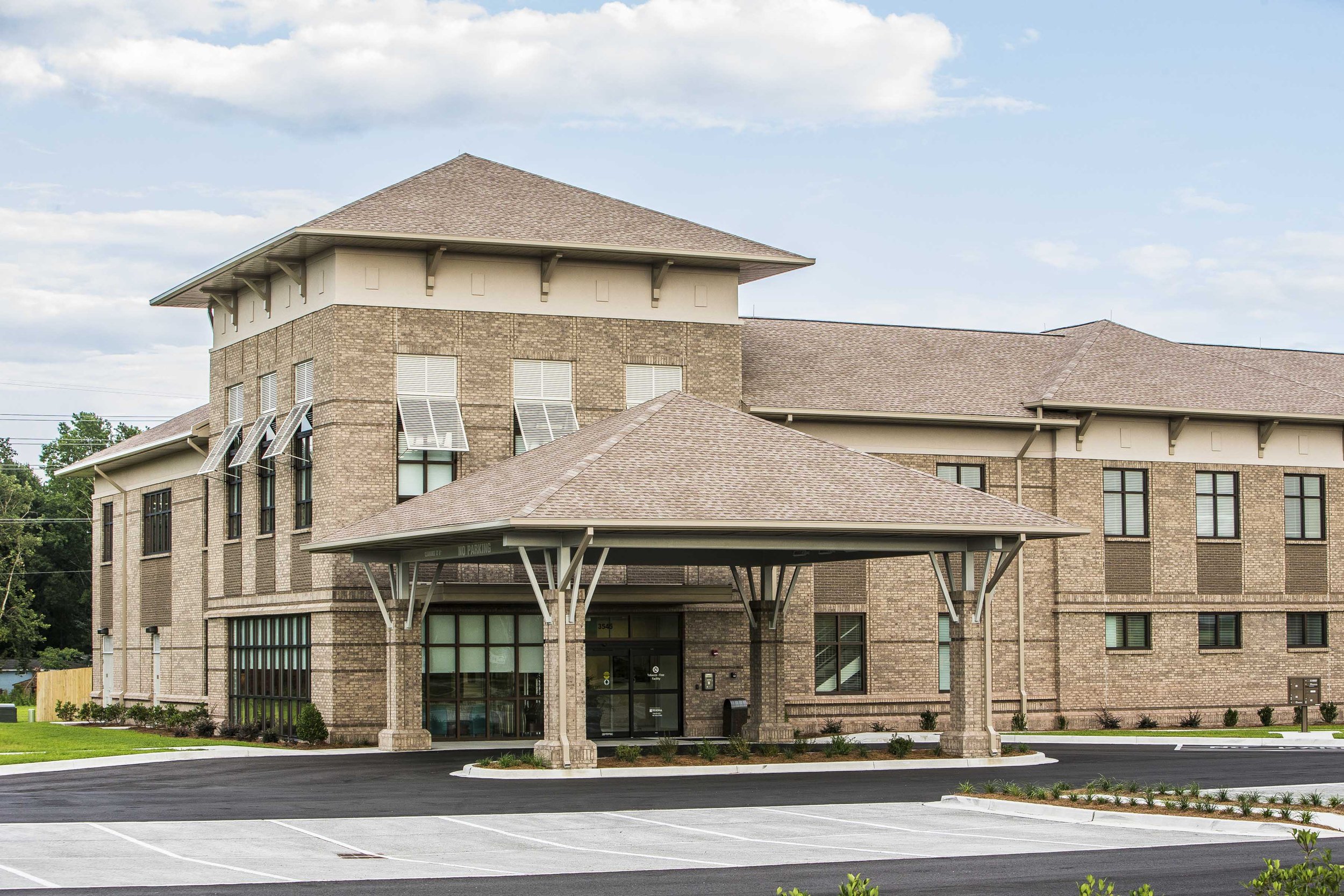 A multi-story brick building with a covered parking area in front, surrounded by a parking lot and grassy areas, under a partly cloudy sky.