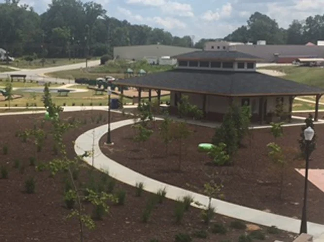 A landscaped park area with a curved walkway, small trees, and a gazebo or covered sitting area with a building in the background.