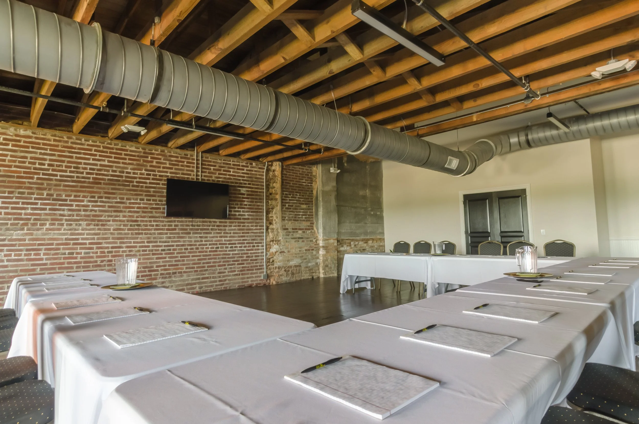 Conference room with tables covered in white tablecloths, set with notepads and pens, and arranged for a meeting or event. Exposed brick wall and wooden ceiling beams, with large ductwork running across the ceiling.