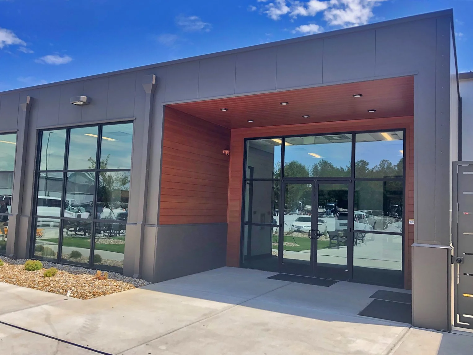 Modern commercial building with a glass front entrance, gray and wood exterior, and a parking lot with cars visible through the windows.