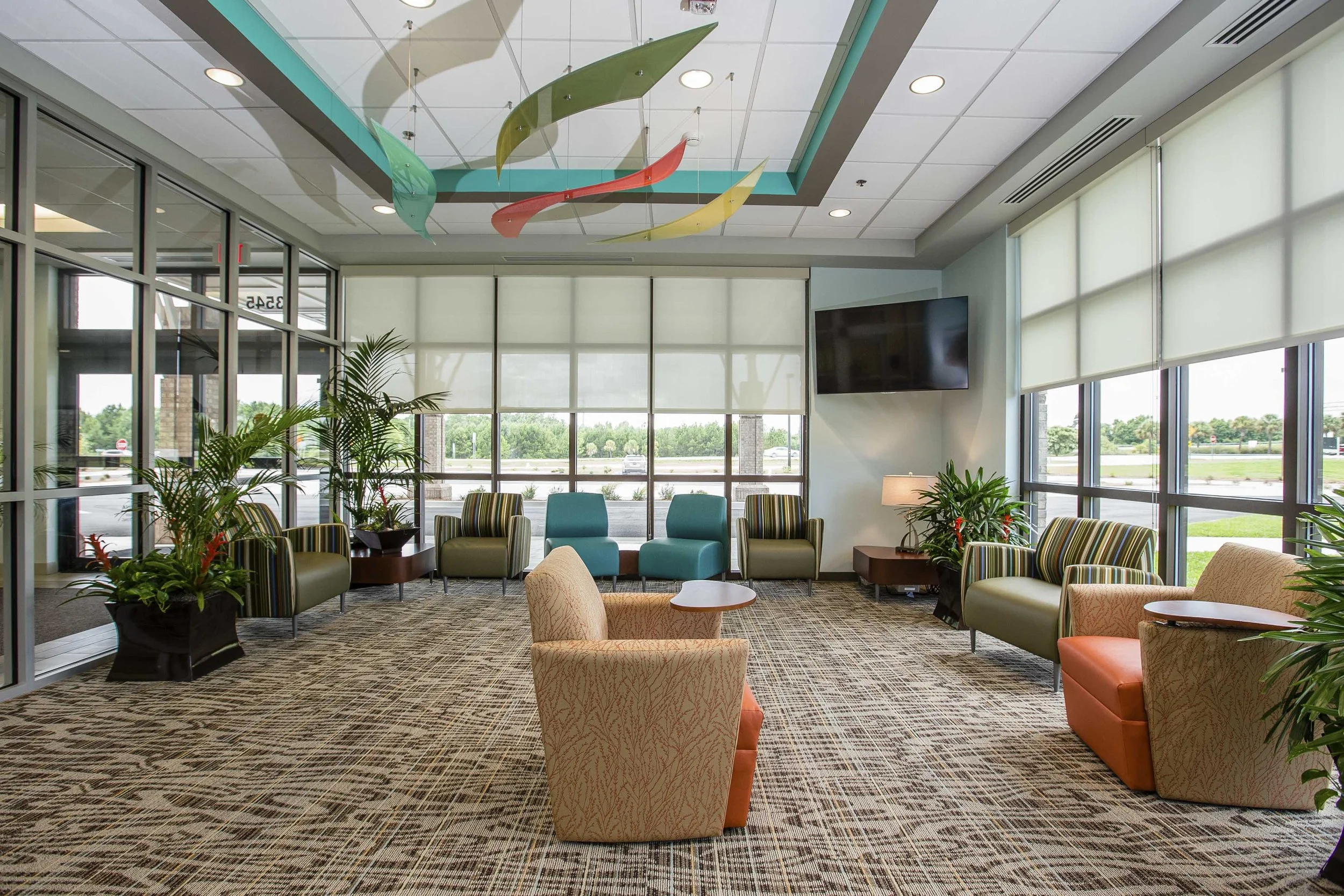 A spacious hotel lobby with large windows, multicolored abstract hanging sculpture, various chairs, and plants.
