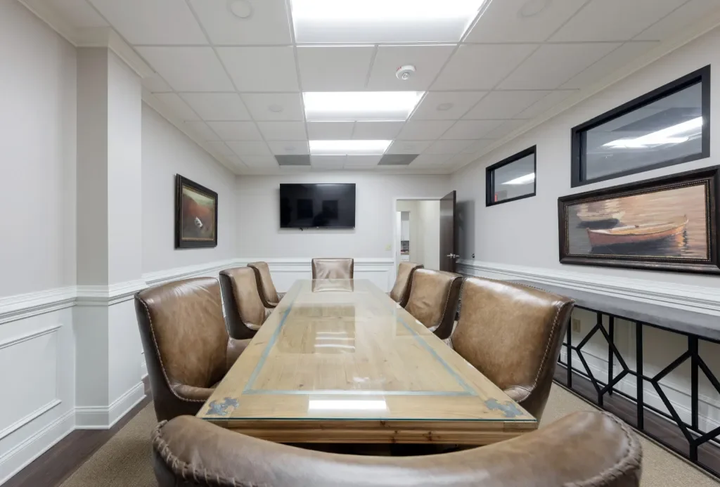 Empty conference room with a long wooden table, leather chairs, a wall-mounted TV, framed paintings, two horizontal windows, and a console table along the wall.