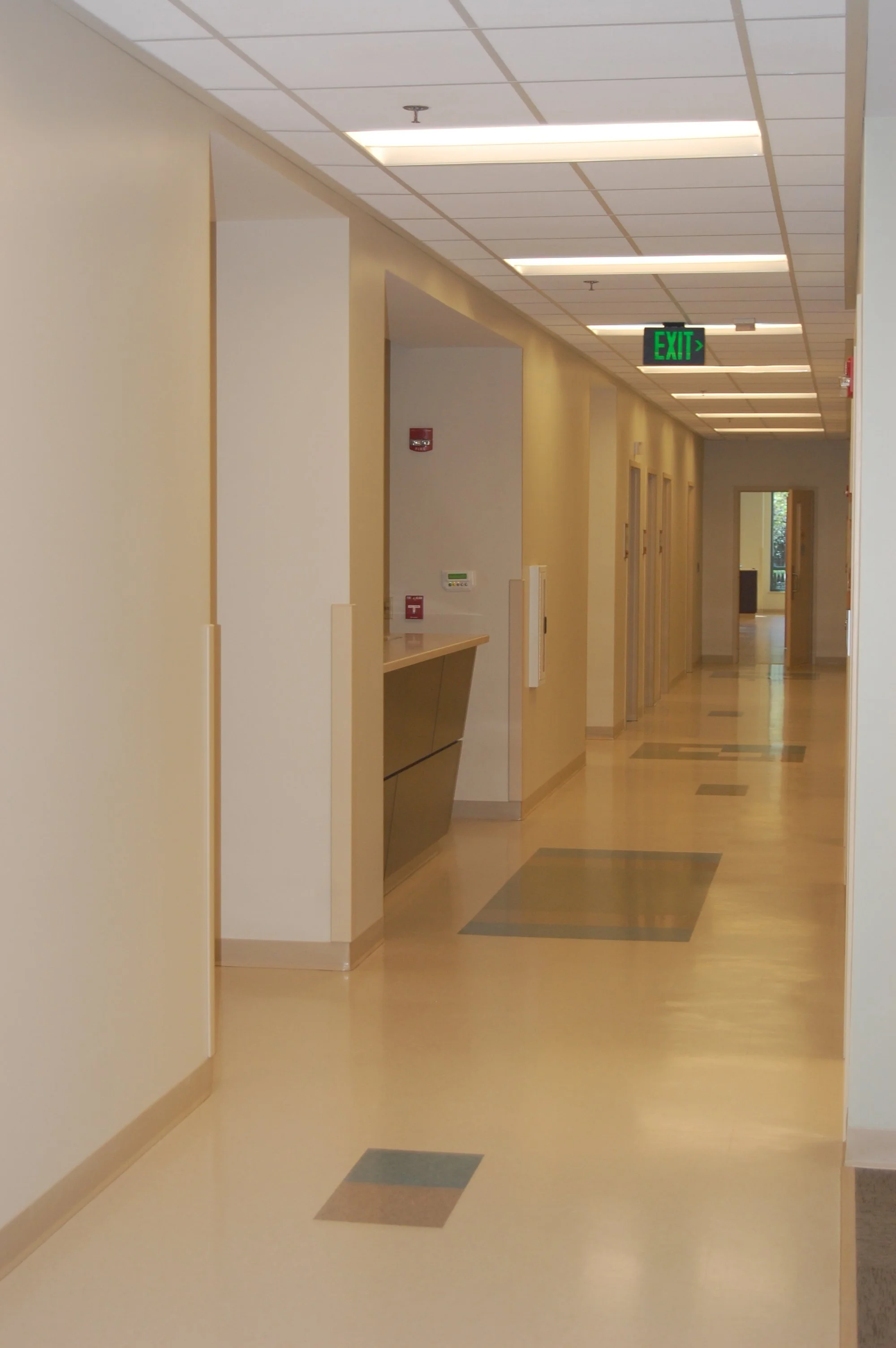 Empty corridor in a building with a beige and yellow color scheme, ceiling lights, and an illuminated green exit sign.