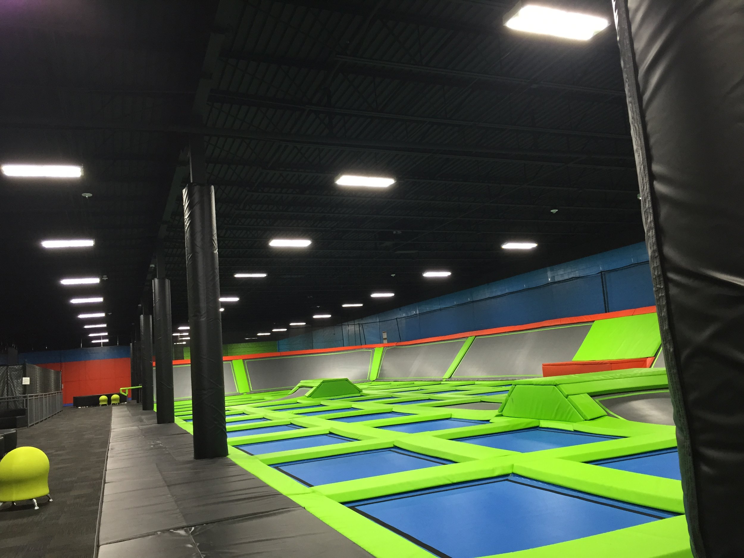 An indoor trampoline park with multiple green and blue trampolines and a padded black wall on the right side.