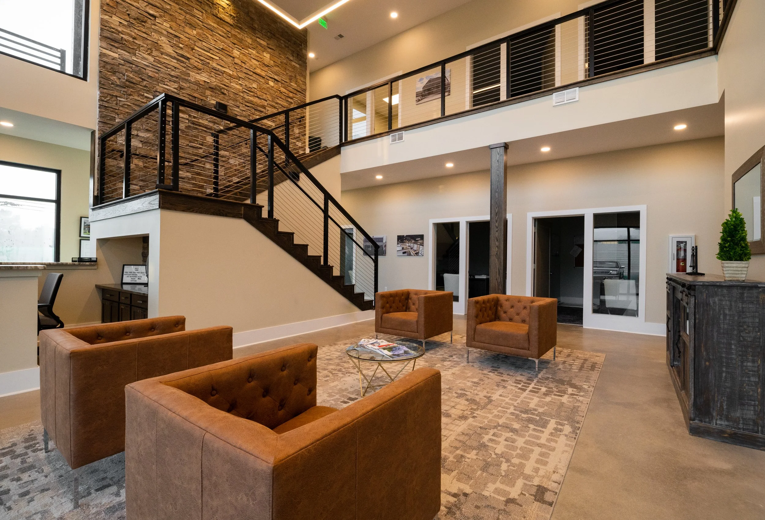 Indoor lobby area with a staircase, multiple cozy brown chairs, a coffee table with magazines, a patterned rug, a fireplace, potted plant, and framed photos on wall, with stone accent wall and modern lighting.