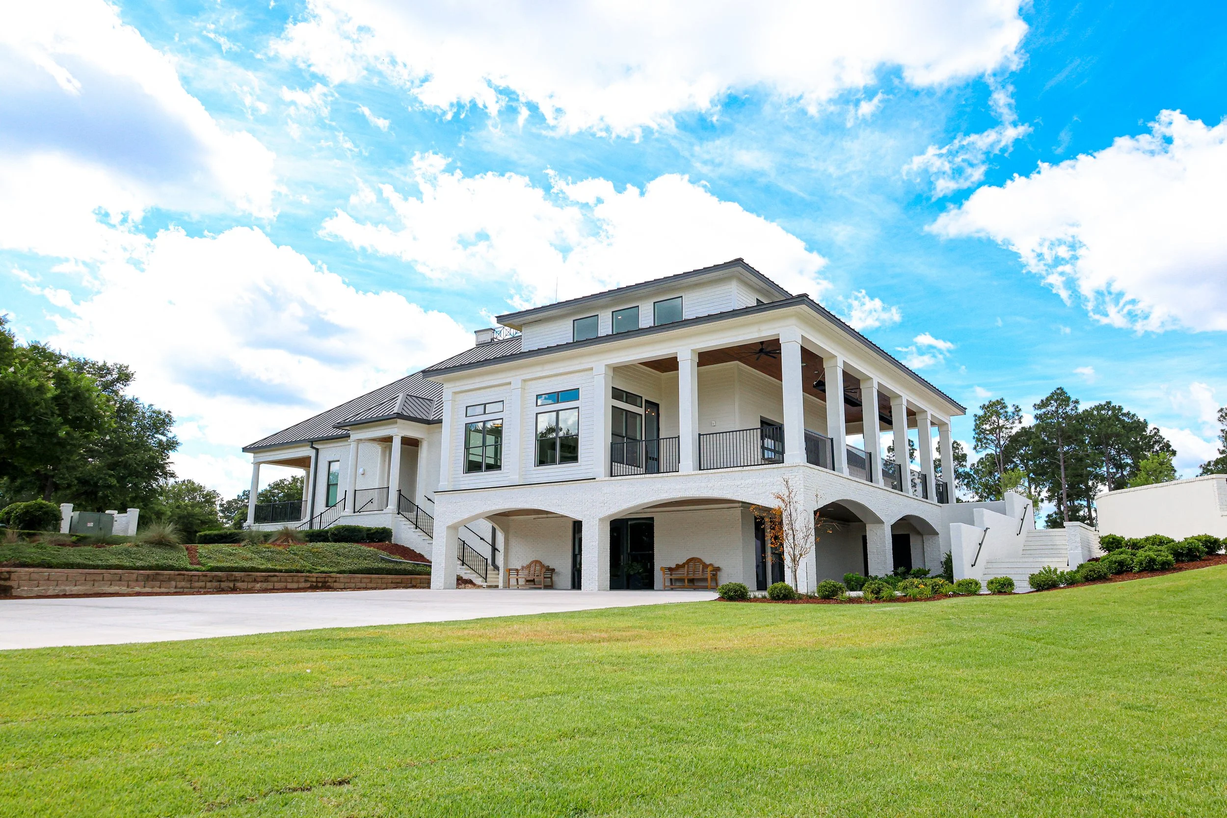 A large, modern, white house with multiple levels and a spacious balcony, situated on a well-maintained lawn with lush green grass, under a blue sky with scattered clouds.