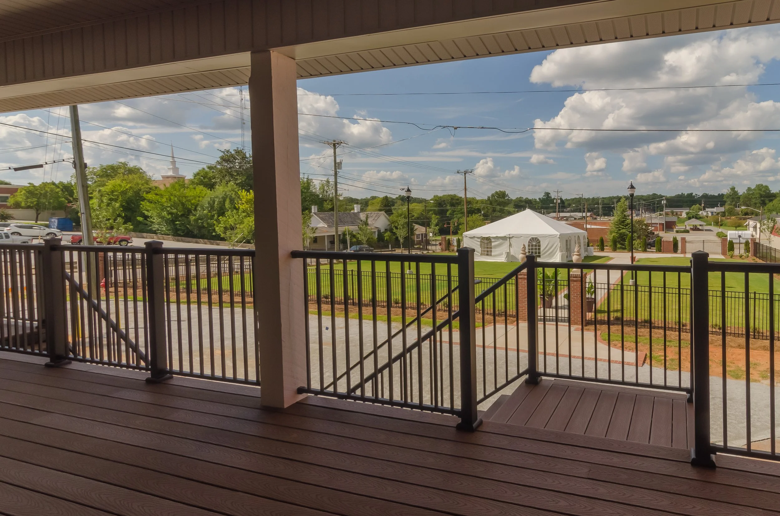 View from a covered wooden porch looking out over a green landscape with trees, a white tent, and houses, under a partly cloudy sky with power lines.