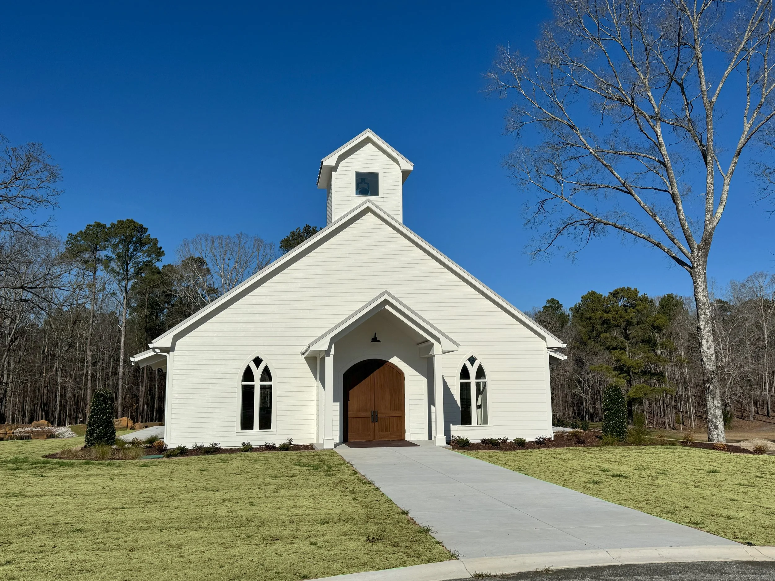 White church with wooden doors, pointed arch windows, and a steeple, surrounded by a lawn with small bushes, under a clear blue sky with leafless and evergreen trees in the background.
