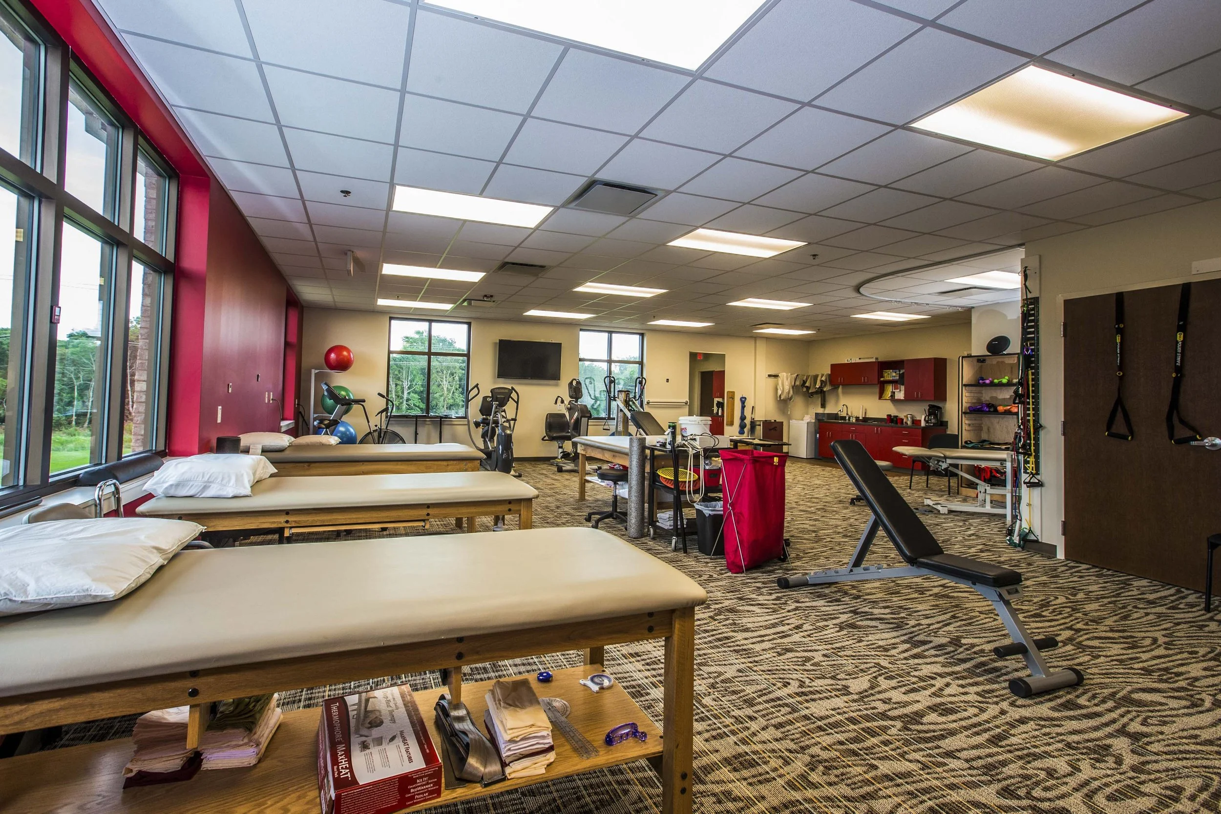 A physical therapy or rehabilitation room with beds, exercise equipment, and medical supplies. Multiple windows provide natural light, and the room has a drop ceiling with fluorescent lights.