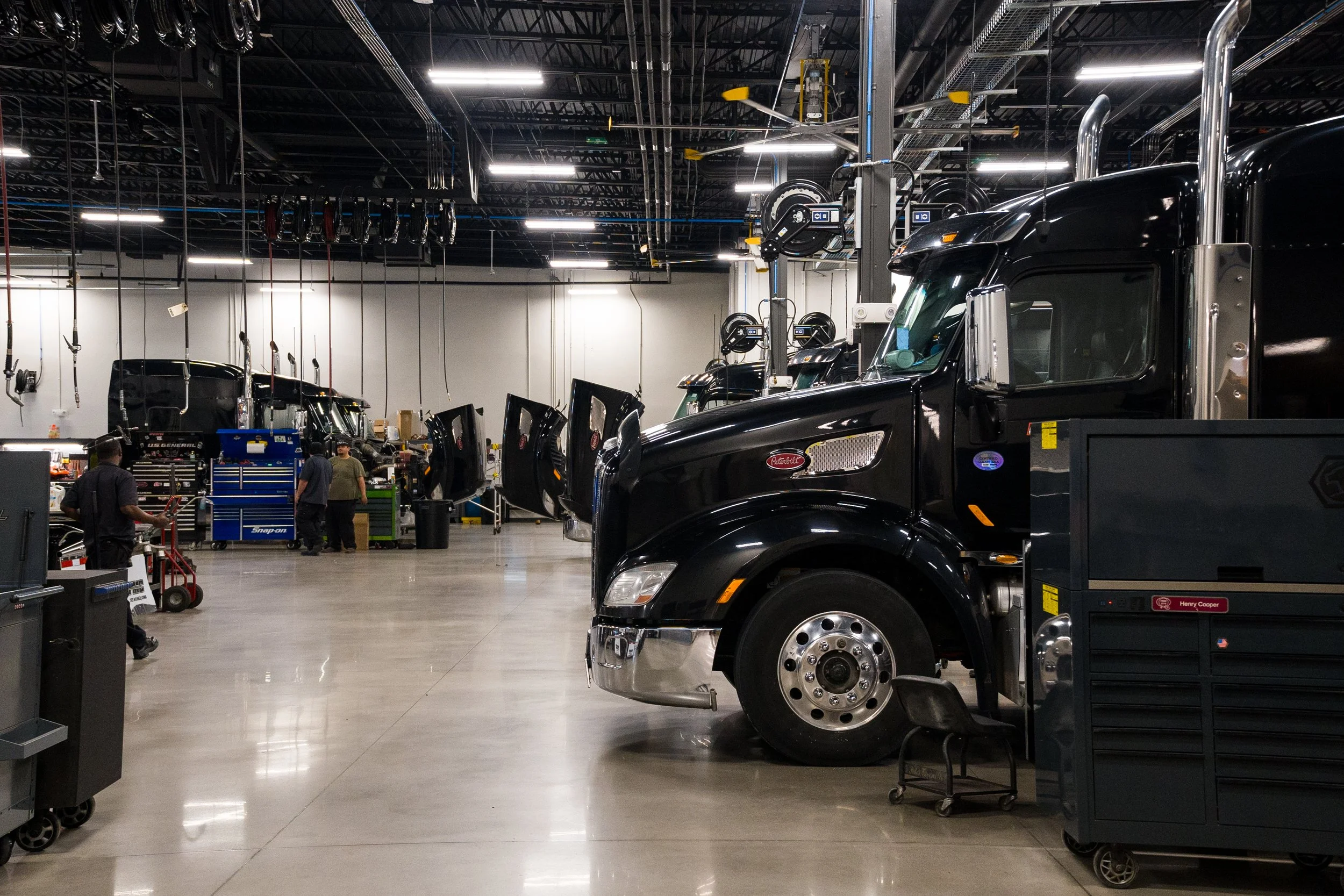 A large indoor facility with multiple trucks parked inside, workstations, and workers engaged in maintenance or repair tasks.