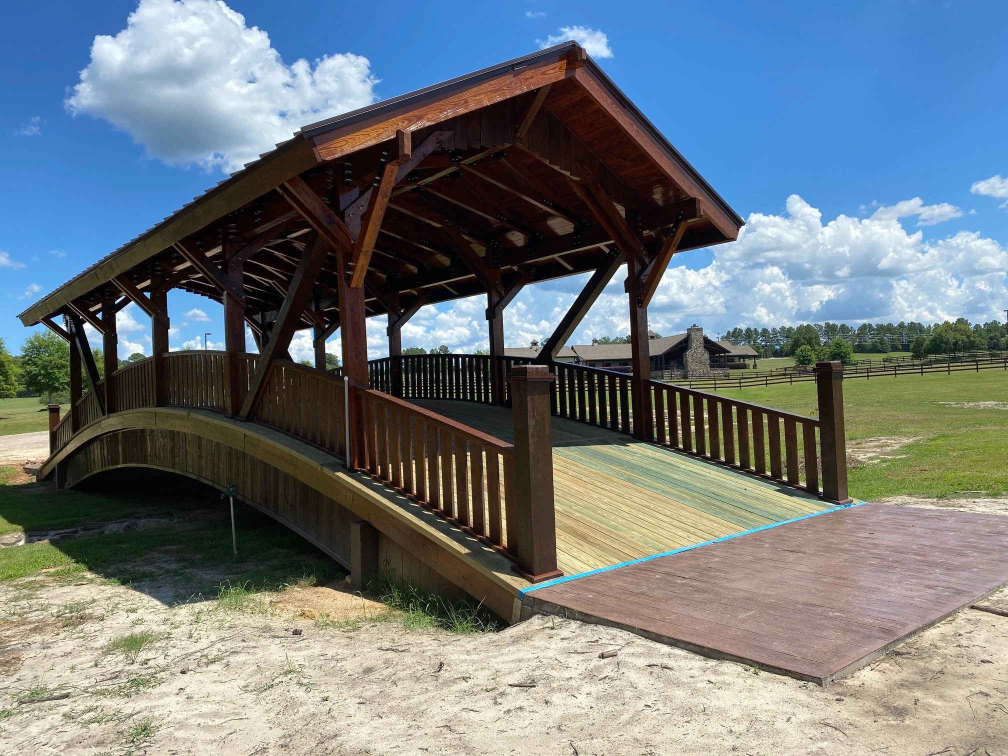 A large wooden bridge with an arched design and a covered roof, situated on a grassy field under a partly cloudy blue sky, with buildings and trees in the background.