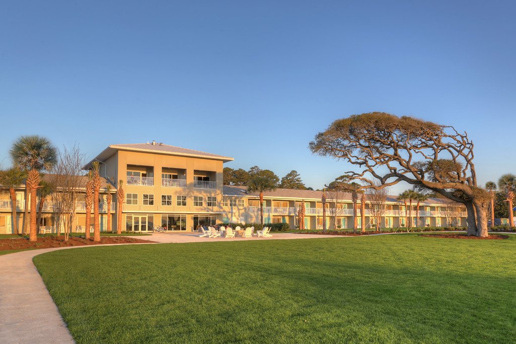 A large, three-story yellow building with balconies, surrounded by palm trees, a well-maintained lawn, and a large, twisted tree, under a clear blue sky.