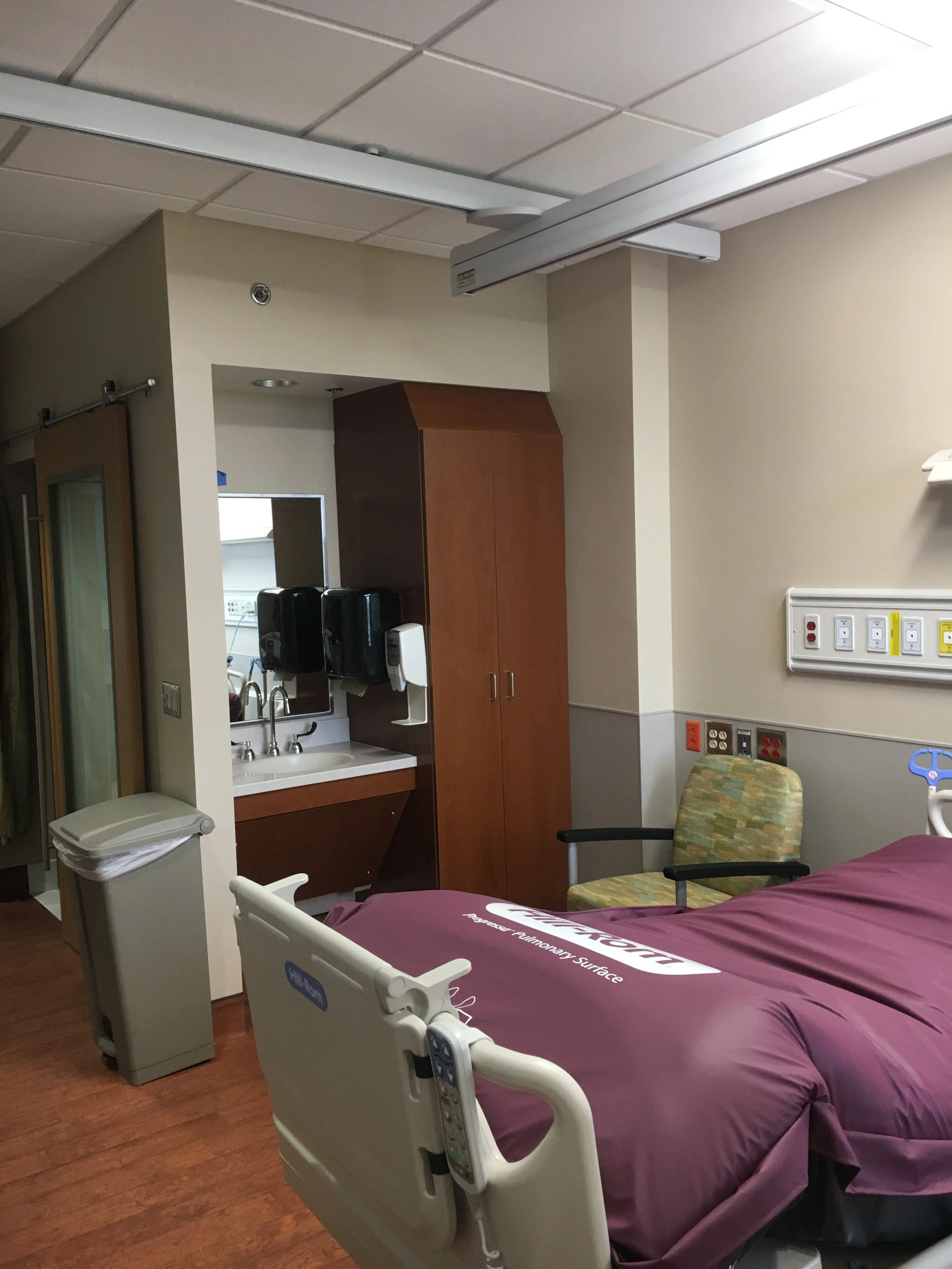 A hospital room featuring a bed with a purple sheet labeled 'Pulmonary Surface', an adjustable IV pole, a yellow-patterned chair, medical equipment including a wall-mounted panel with outlets and switches, a sink with hand sanitizers, and a wooden ca