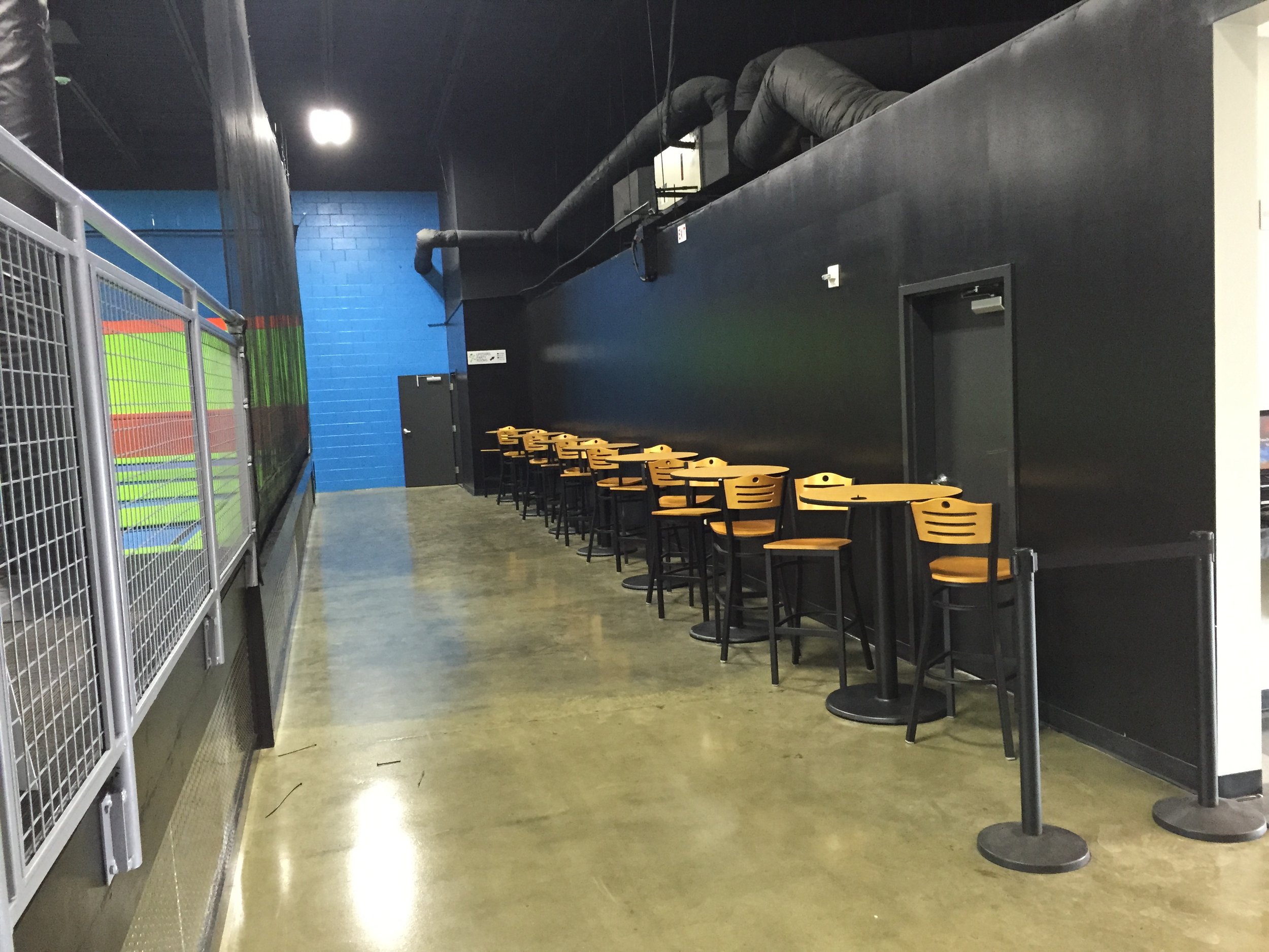 Indoor space with black walls and ceiling, black ventilation ducts along the ceiling, a row of wooden chairs with black metal legs lined against the wall, and a closed black door. The floor is polished concrete.