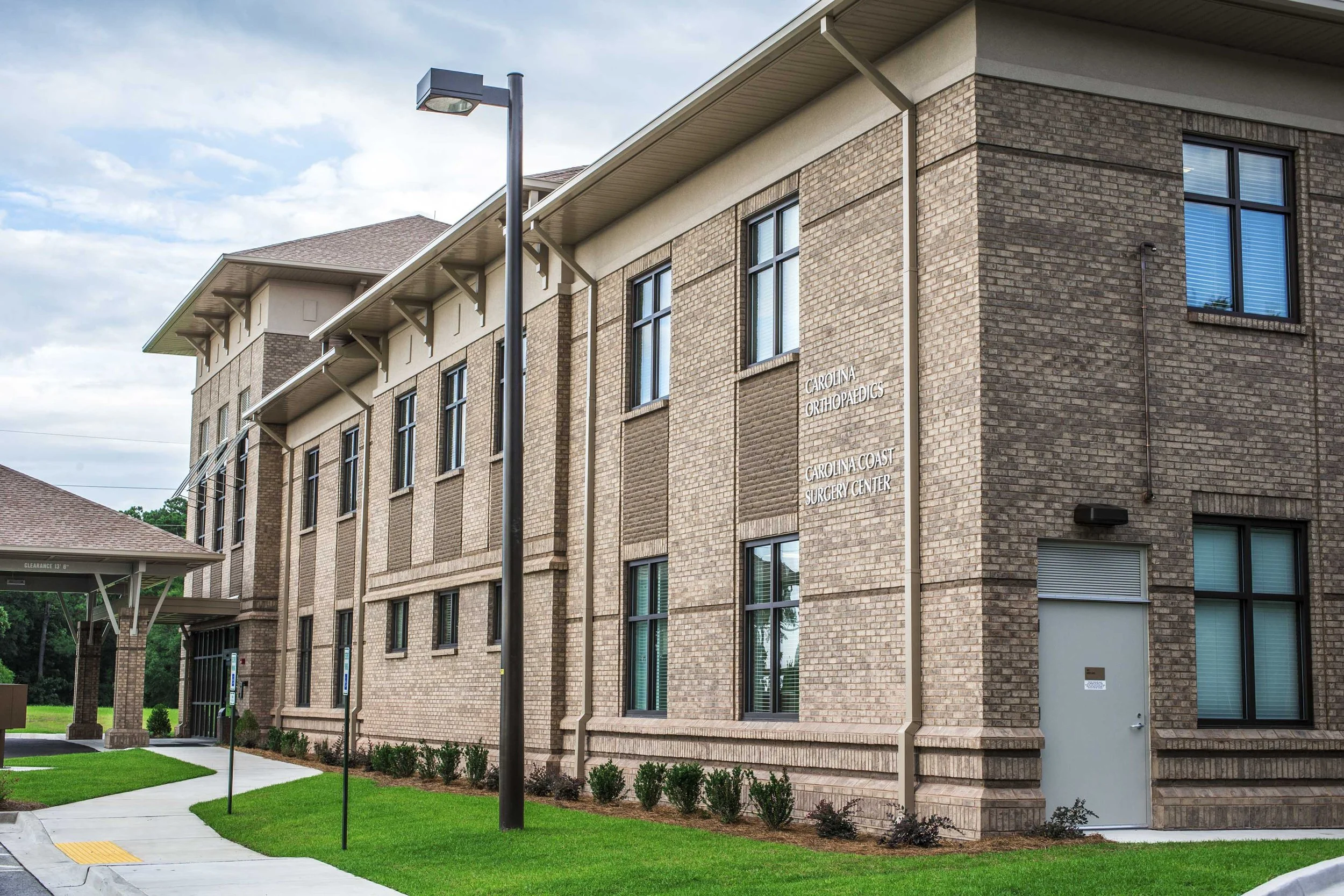 Exterior of a brick medical building with signs indicating it is the Carolina Orthopaedics and Carolina Coast Surgery Center. There is a sidewalk with a textured tactile paving strip, green grass, small bushes, and a streetlamp.